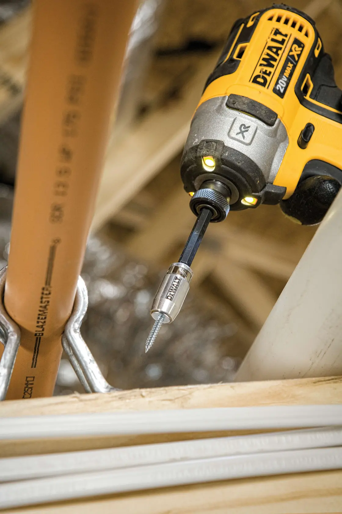 A DEWALT cordless power drill is shown driving a screw into a metal bracket attached to a plastic pipe. The drill has a DEWALT branded bit and is being used in a construction or plumbing setting.