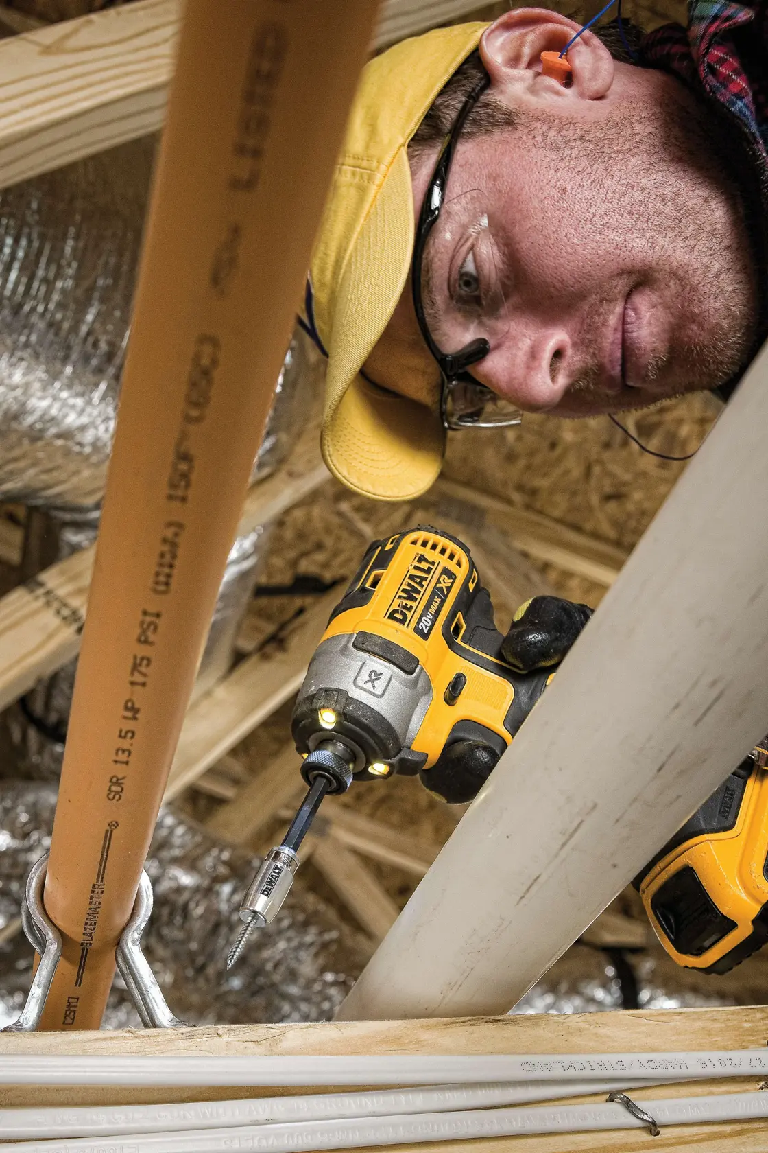 Close-up of a DEWALT cordless impact driver being used to fasten a screw into a wooden beam during construction, with PVC pipes and pipe hangers visible in the foreground.