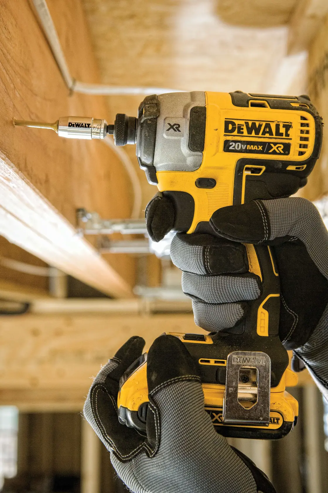 A close-up view of a person wearing work gloves using a DEWALT 20V MAX XR cordless impact driver to drive a screw into a wooden beam indoors. The tool is yellow and black.