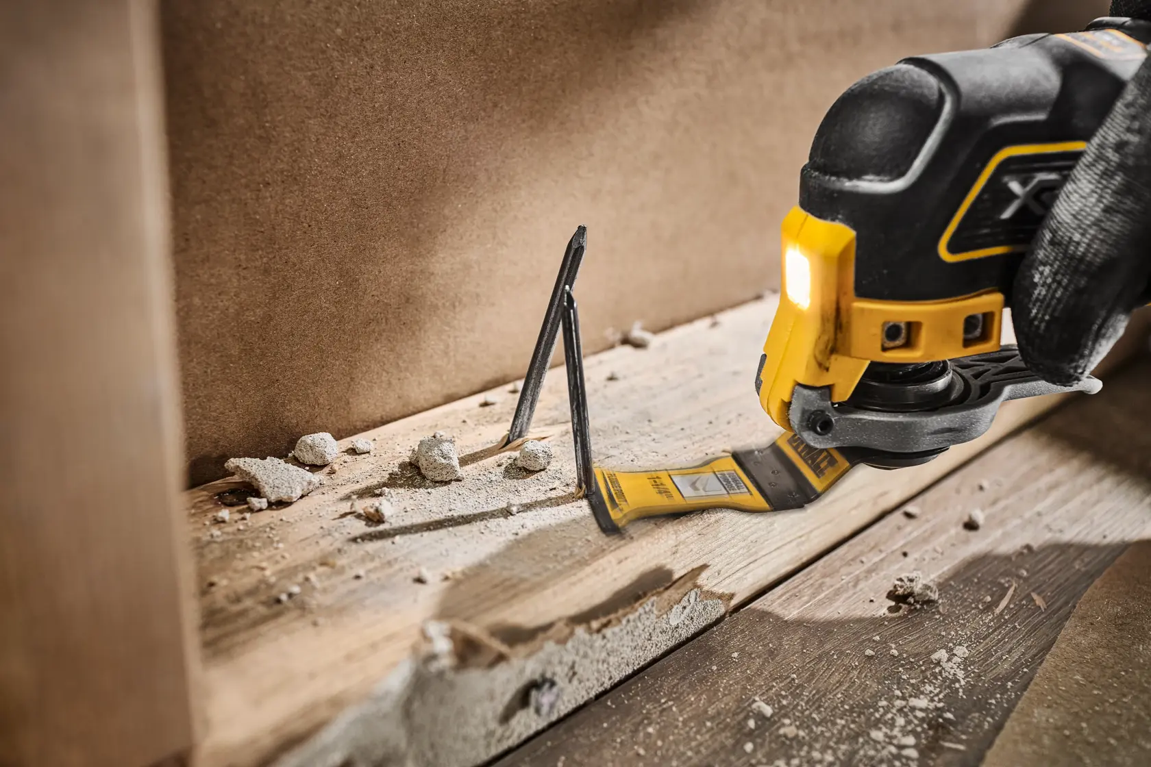 A DEWALT oscillating multi-tool with a blade attachment is being used to cut through a wooden board with embedded nails. There is construction debris scattered around the work area.
