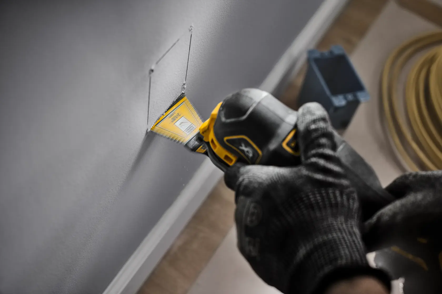 A close-up of a person wearing gloves, using a DEWALT oscillating multi-tool with a yellow blade attachment to cut a rectangular opening in a wall. The tool appears to be cutting drywall, and electrical wiring and a blue outlet box are visible in the background.