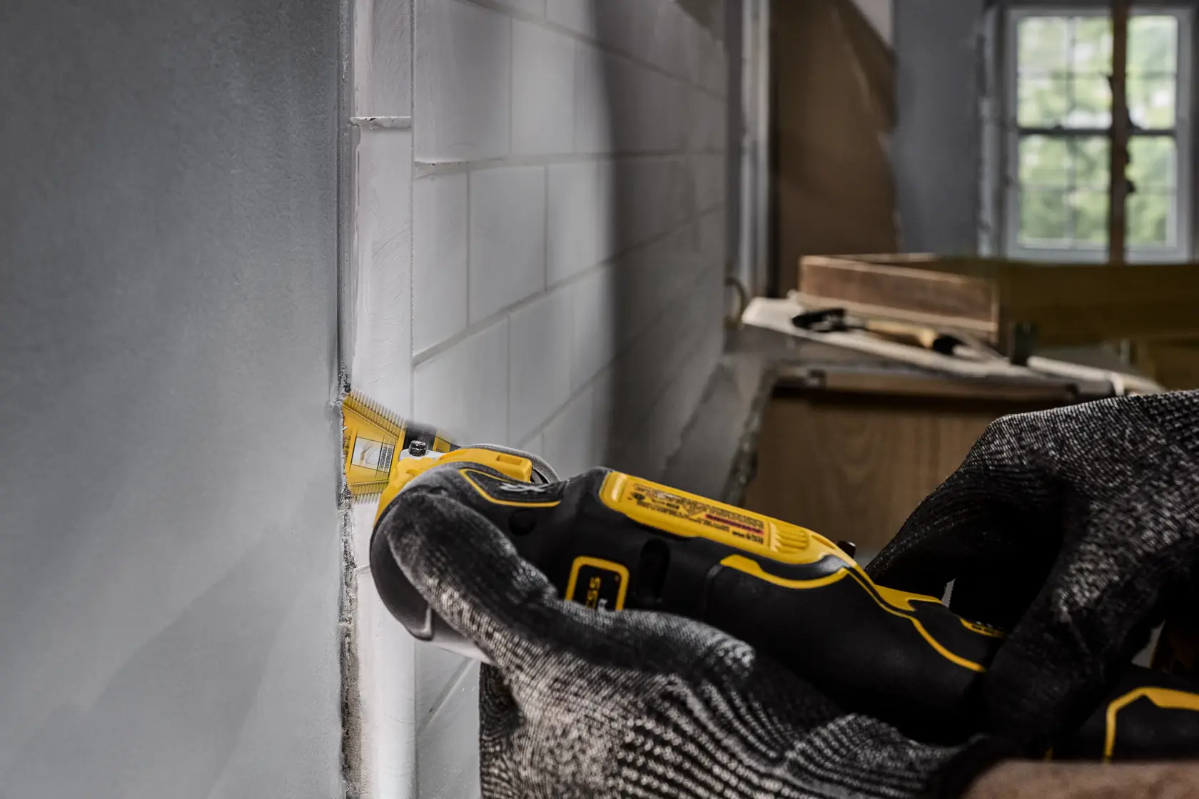 A person wearing protective gloves uses a DEWALT oscillating tool to cut or trim around white wall tiles during renovation or construction work.