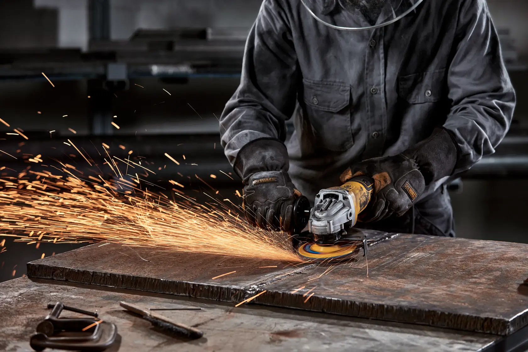 A person wearing protective gloves and clothing uses a DEWALT angle grinder to cut or grind a metal sheet, producing bright sparks in a workshop setting.