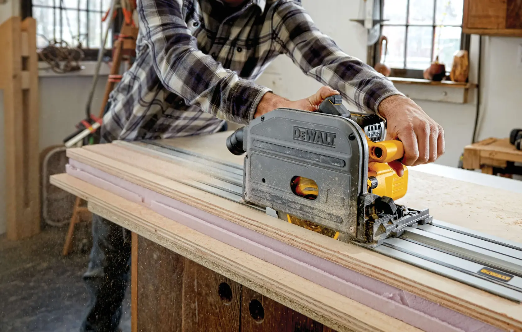 A person using a DEWALT circular saw on a guide rail to cut a sheet of plywood in a workshop.