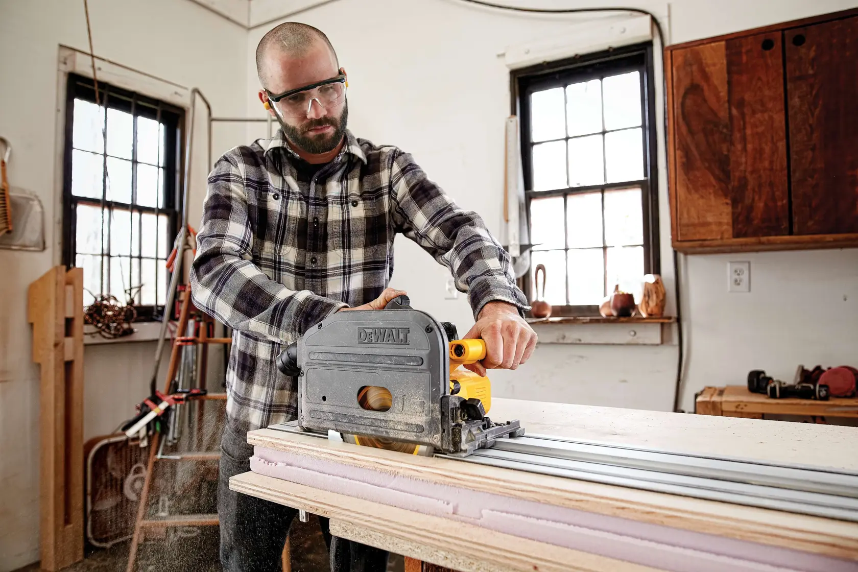 A person using a DEWALT circular saw with a guide rail to cut a sheet of wood in a workshop. The person's face is blurred. There are tools and wooden objects visible in the background.