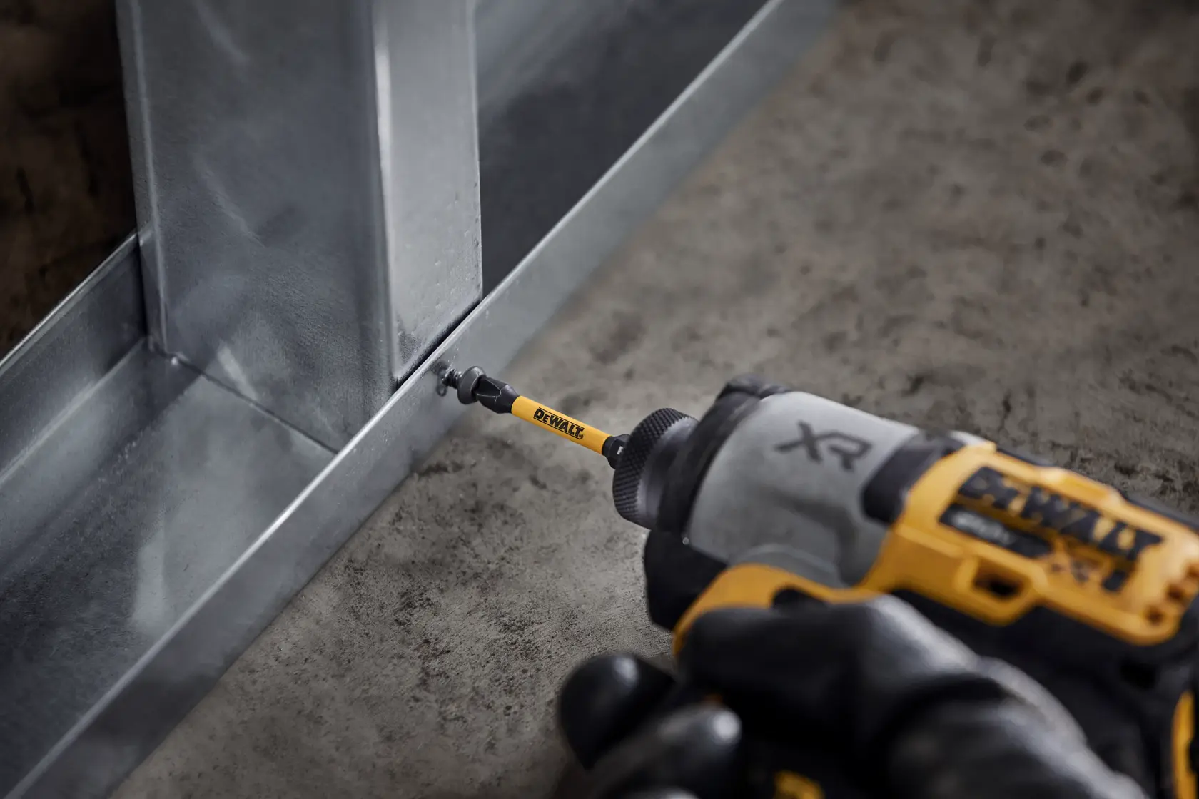 A close-up image of a DEWALT impact driver being used to fasten a screw into a metal frame on a concrete surface. The DEWALT branded bit is visible in action.