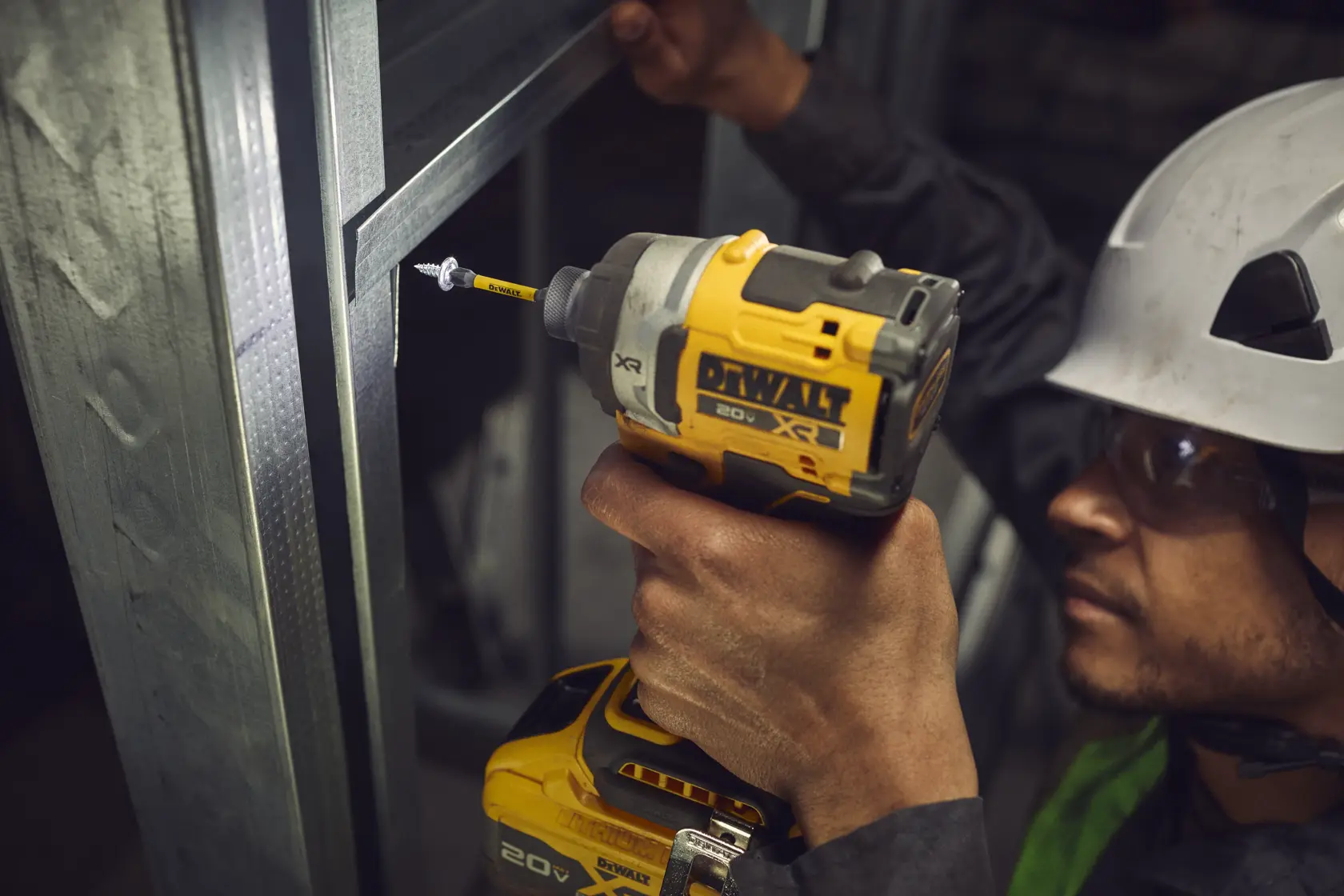 A close-up of a person using a yellow DEWALT cordless drill to drive a screw into a metal frame. The person's hand is holding the drill, and they are wearing a safety helmet. The DEWALT branding is visible on the drill.
