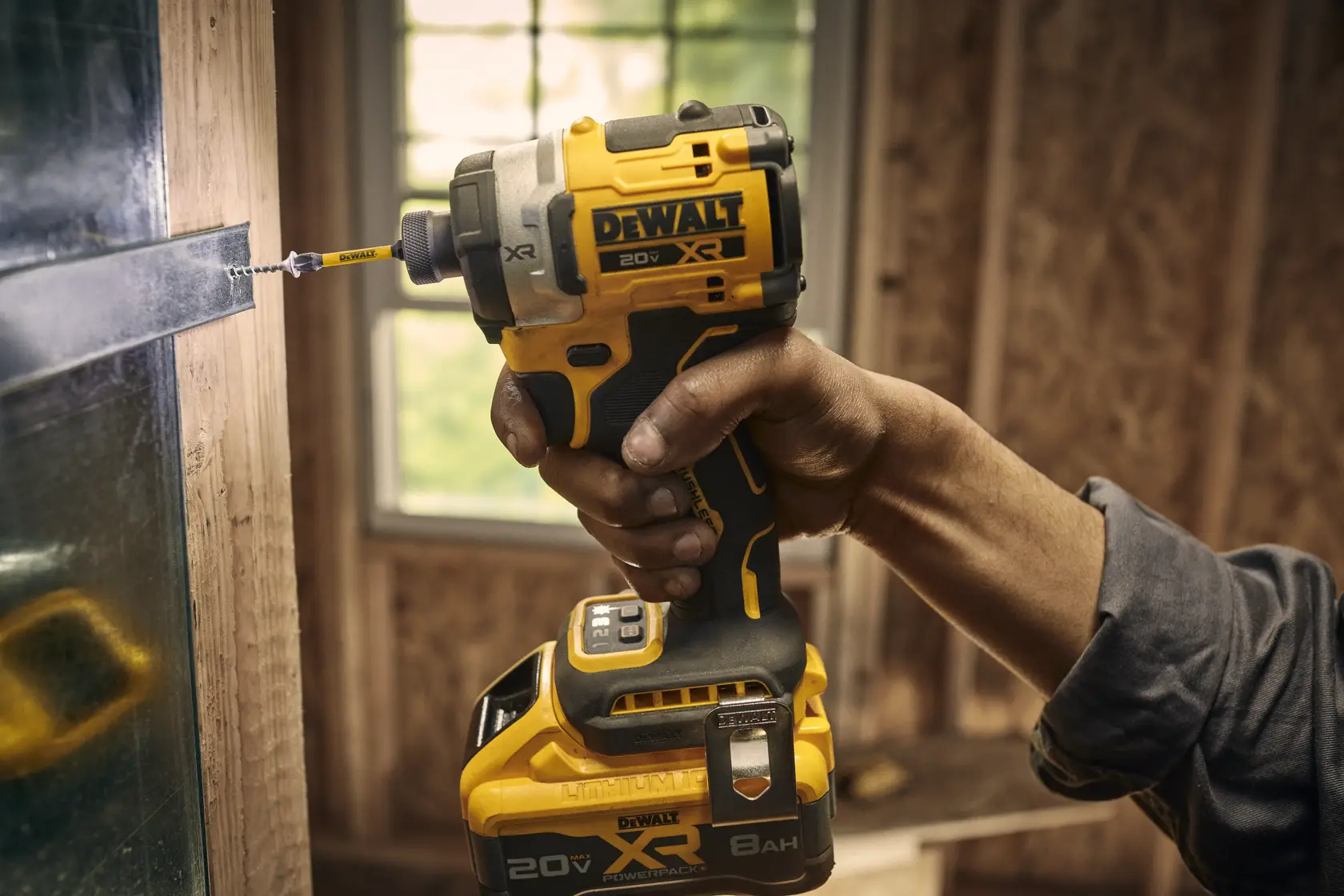 A hand holds a DEWALT 20V XR cordless impact driver, driving a screw into a metal surface inside a construction setting with wooden walls and a window in the background.
