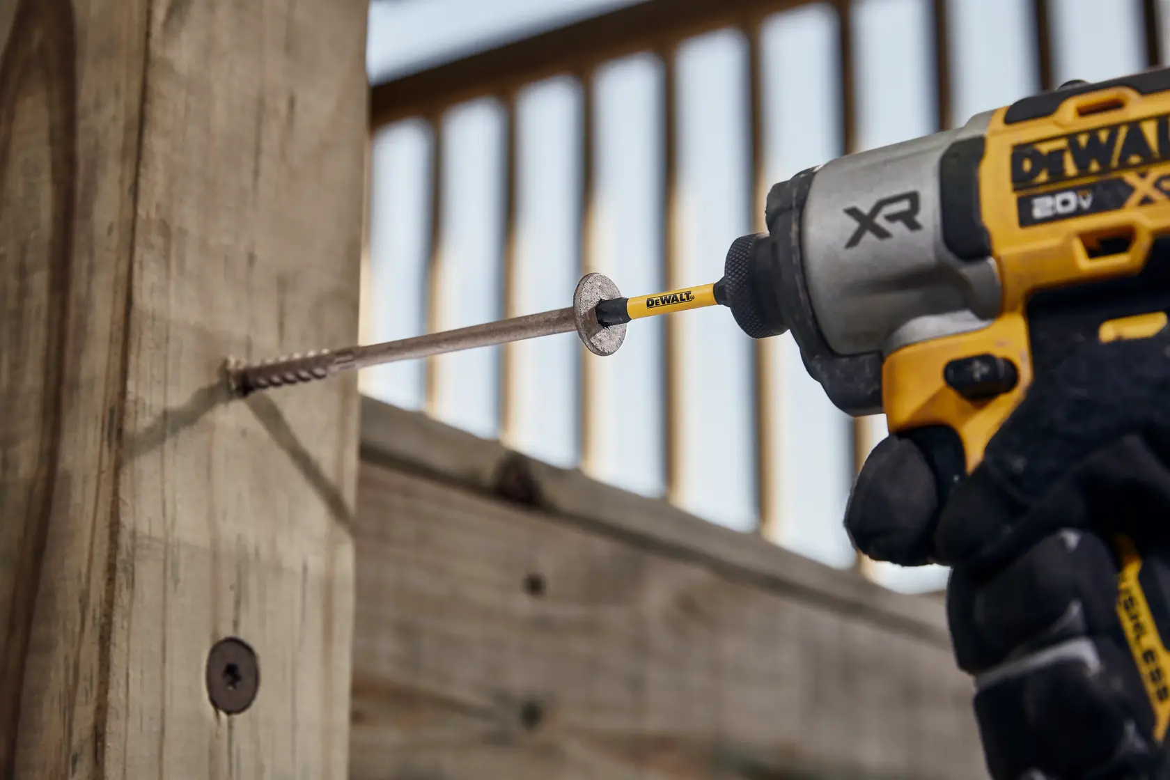 A close-up of a DEWALT power drill driving a long screw into a wooden plank. The worker is wearing gloves and the DEWALT branding is visible on the drill and driver bit.