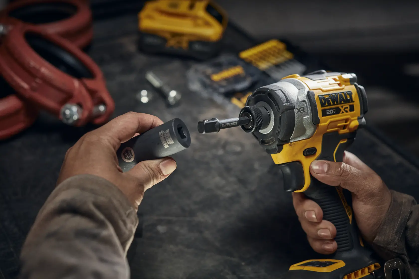 Close-up of hands holding a DEWALT cordless impact driver and an adapter socket in a workshop setting, with various tools and parts visible in the background.