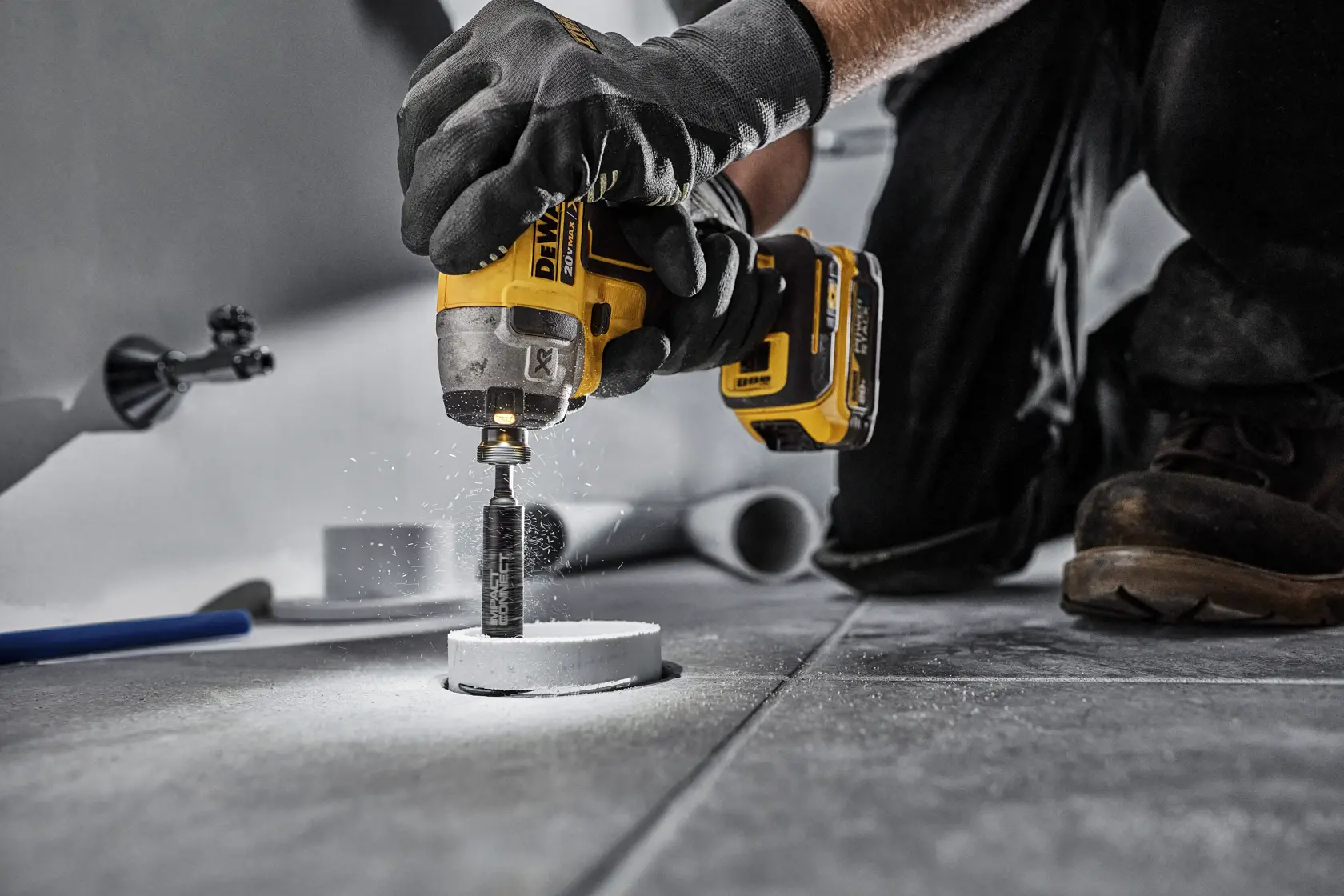 A person using a DEWALT cordless impact driver with a hole saw attachment to drill into a tile floor. The individual is wearing work gloves and boots, and there are dust particles visible during the drilling process.
