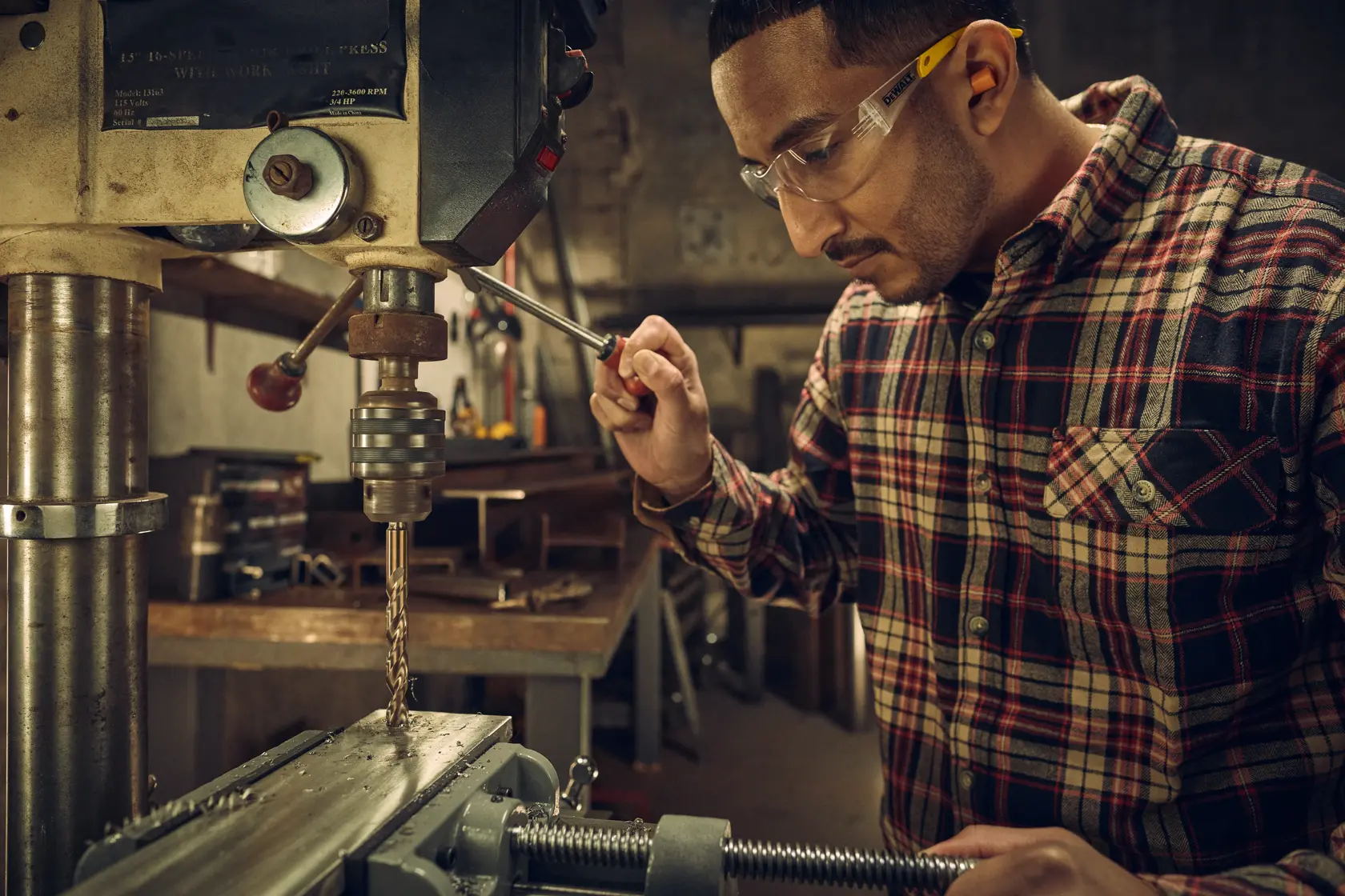 A person wearing a plaid shirt operates a bench drill press in a workshop, with the person's face blurred for privacy. Metalworking equipment and tools are visible in the background.