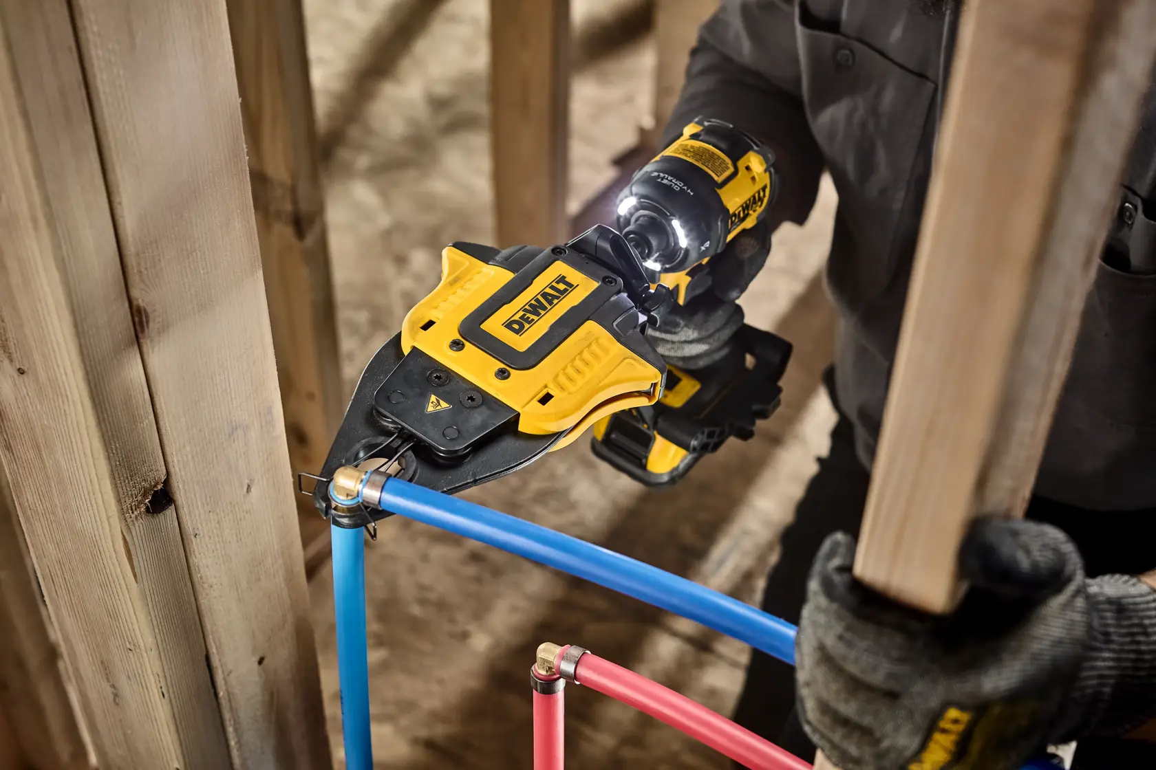 A DEWALT power tool being used to crimp a fitting onto a blue pipe in a construction setting, with gloves and wooden framing visible.