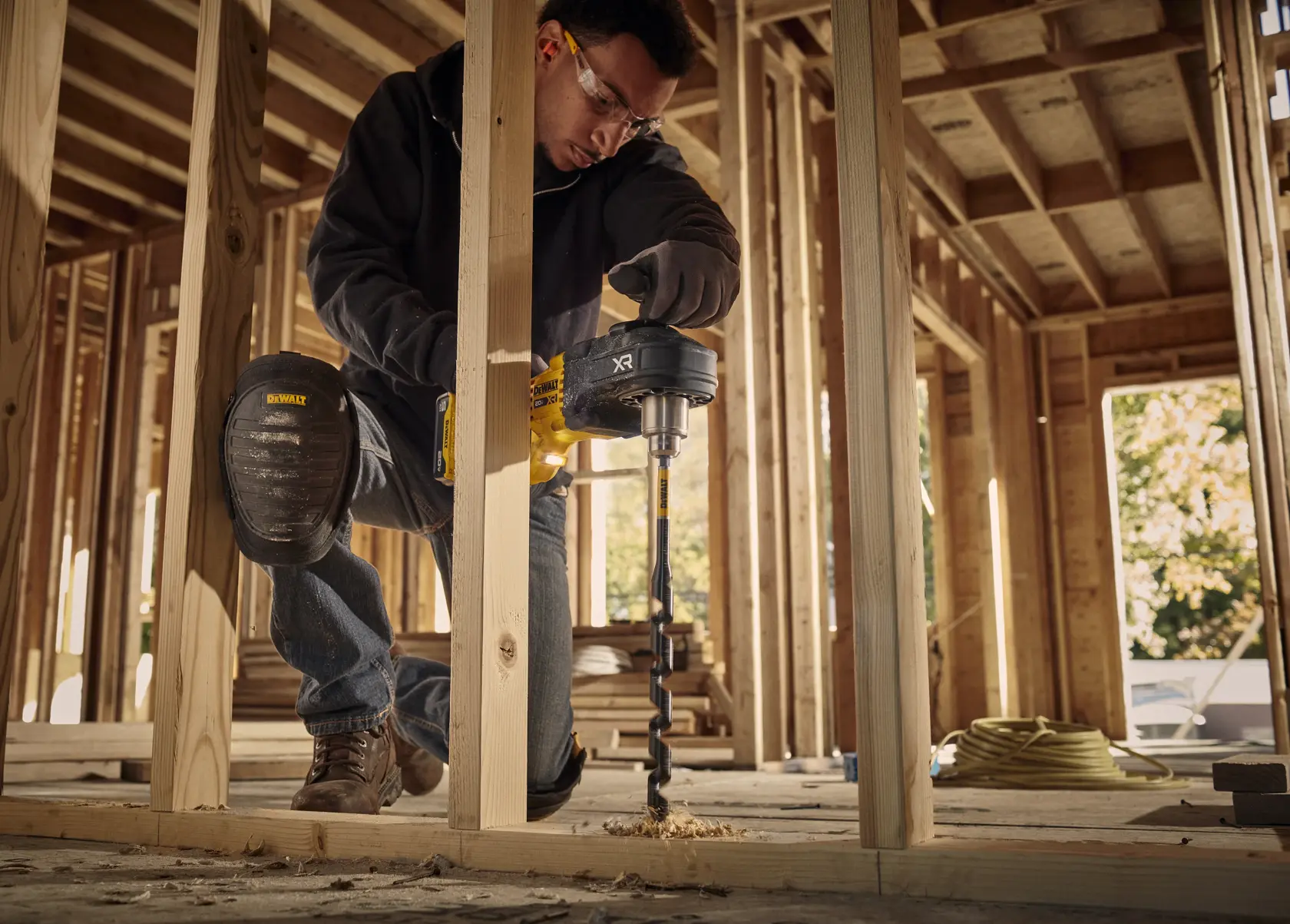 A person wearing a DEWALT knee pad is using a DEWALT power drill with a large auger bit to drill through a wooden beam in a construction site with unfinished framing.