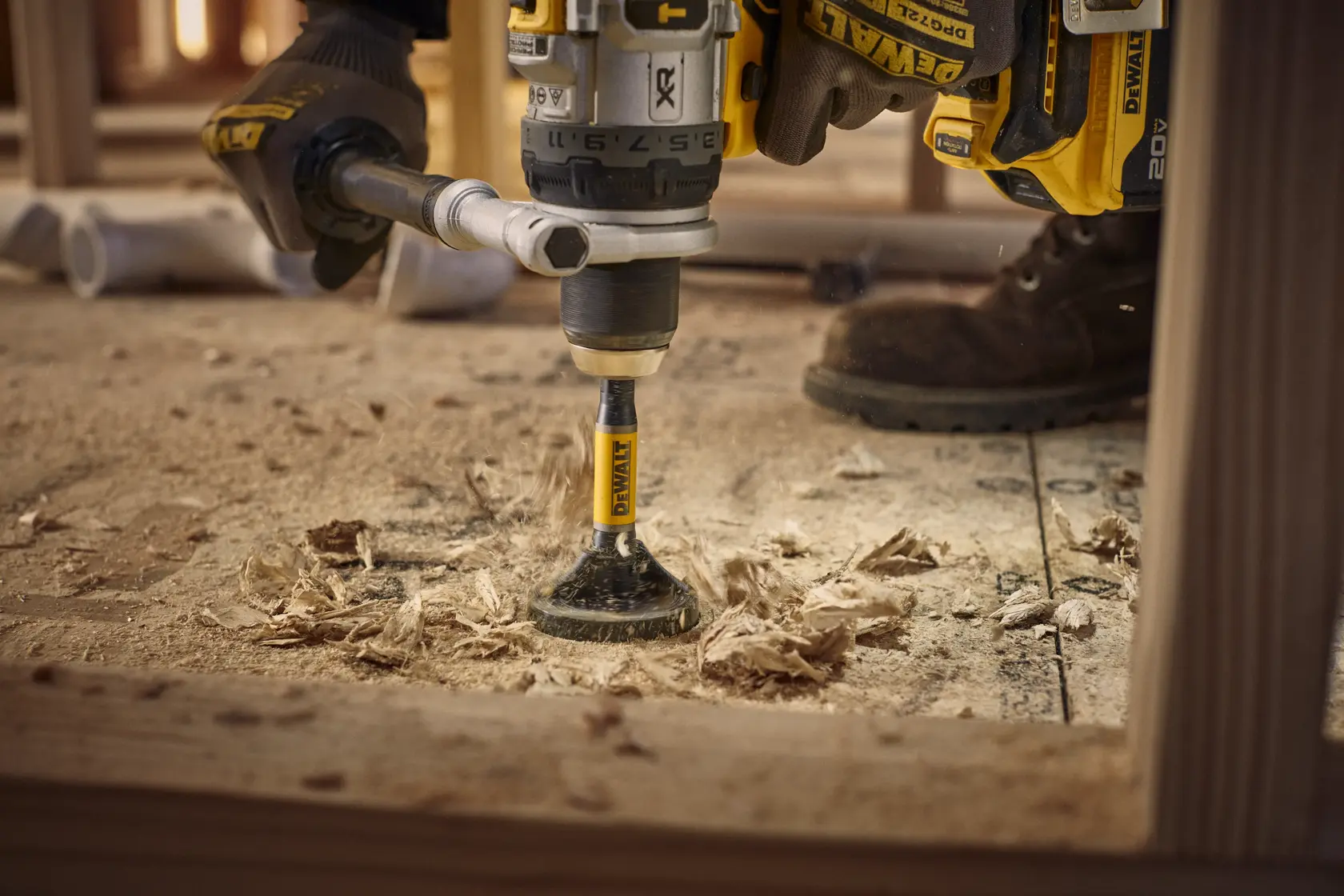 A close-up view of a DEWALT drill with an auger bit being used to bore a hole in a wooden surface, with wood shavings scattered around and a worker wearing gloves and boots.