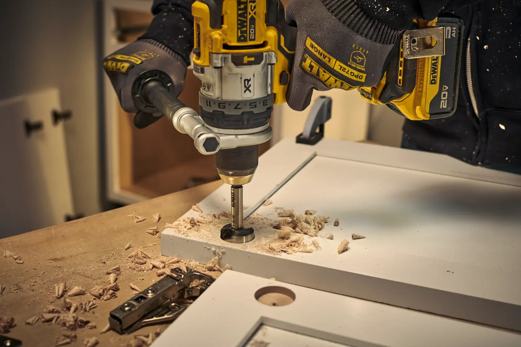 A person wearing DEWALT gloves is using a DEWALT cordless drill with a Forstner bit to drill a hole in a white cabinet door, with wood shavings scattered on the workbench.