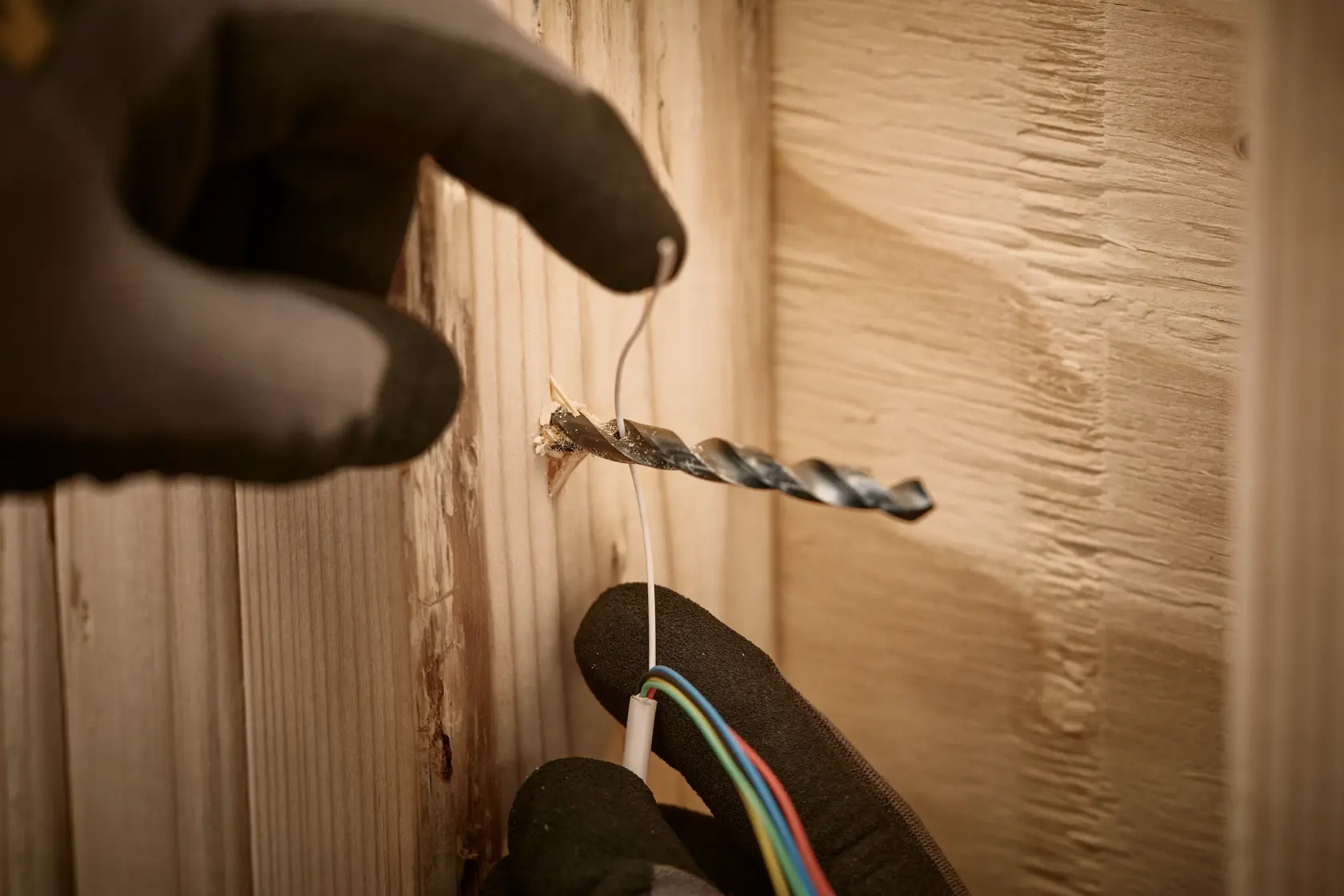Close-up of a gloved hand pushing electrical wires through a hole in a wooden surface next to a drill bit.