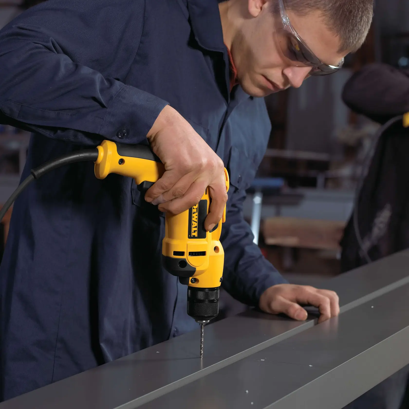A person in a blue work jacket is using a yellow electric drill to make a hole in a metal beam in a workshop setting.