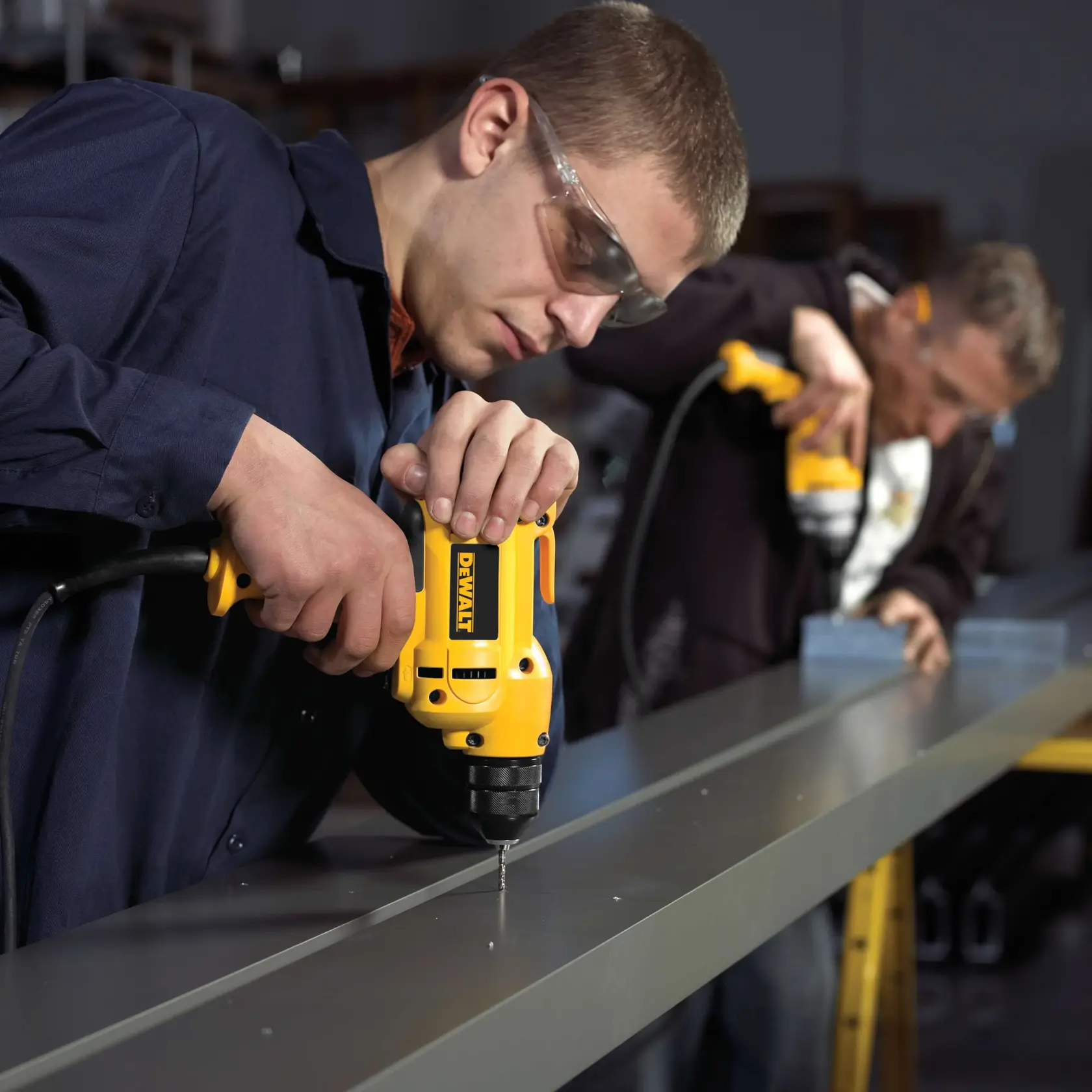 Two people using yellow DEWALT electric drills to make holes in a long metal beam in a workshop setting. Both individuals are wearing protective eyewear and working side by side.