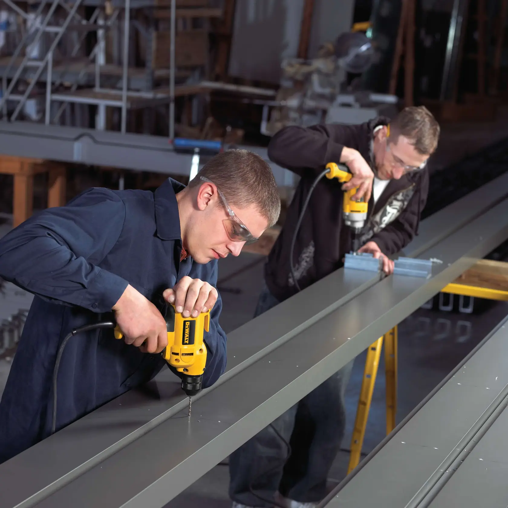 Two people using DEWALT corded electric drills to work on metal beams in an industrial workshop. Their faces are blurred for privacy. The setting includes metal structures and ladders in the background.