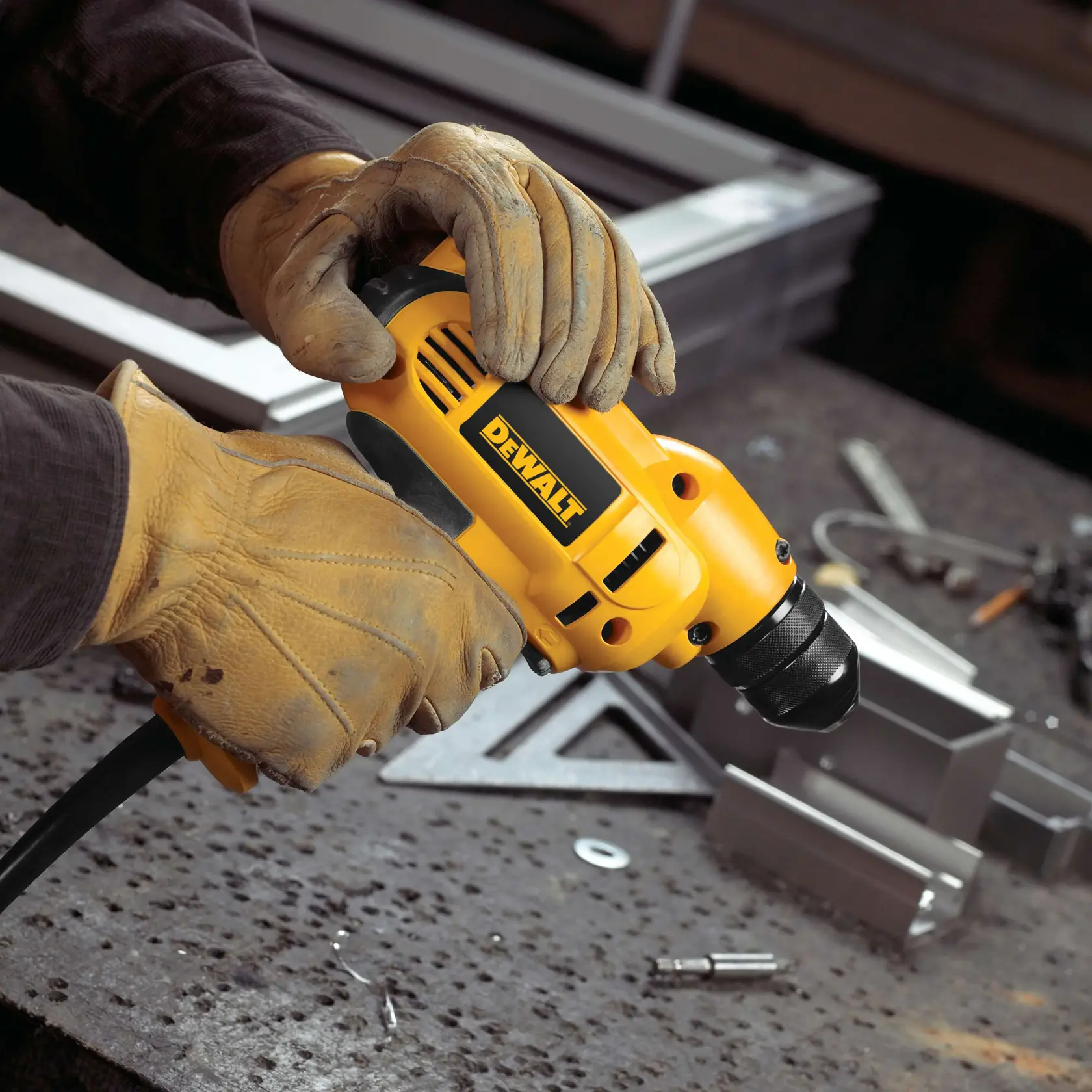A person wearing brown work gloves is holding a yellow DEWALT corded drill on a metal workbench with various tools and metal parts scattered around.