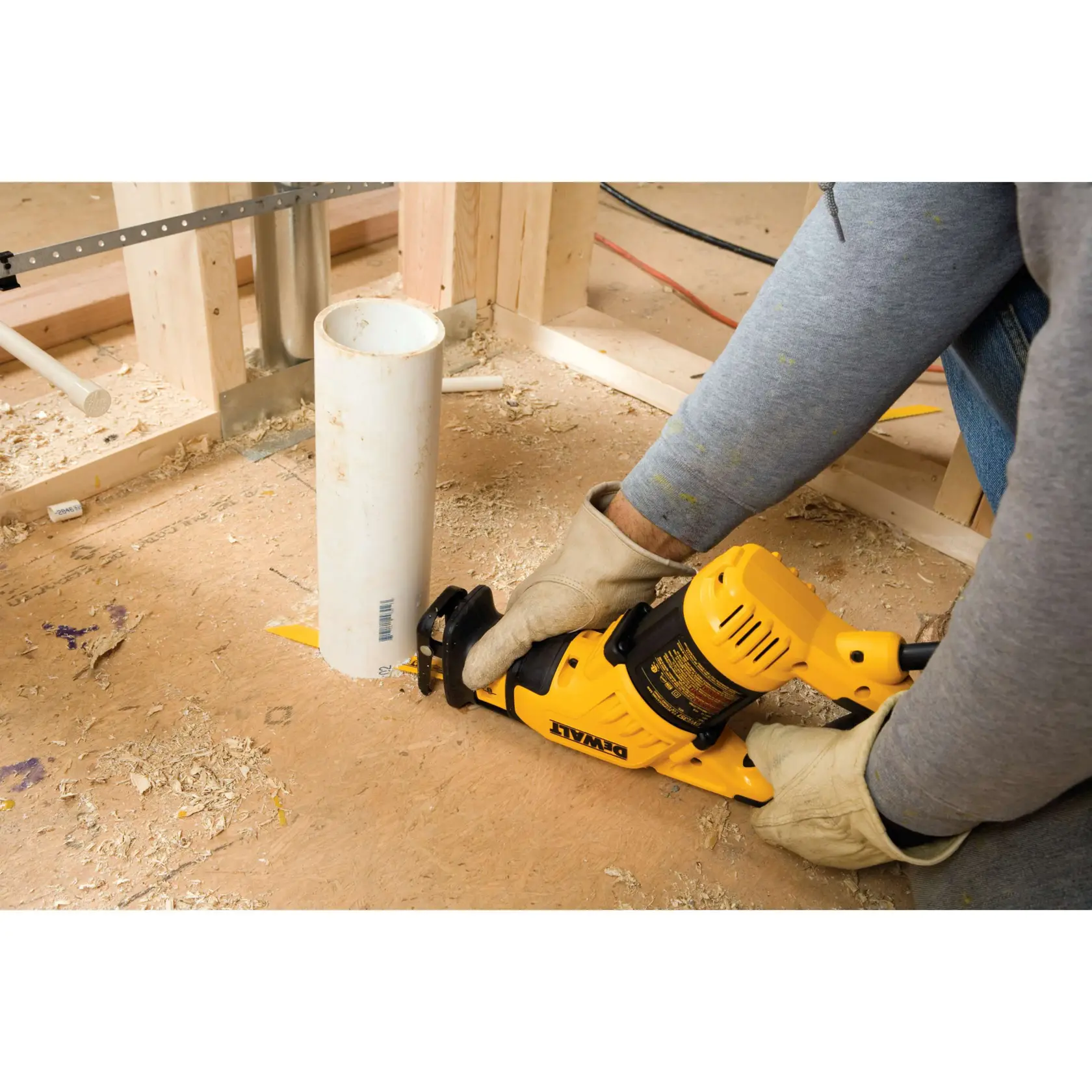 A person wearing gloves uses a DEWALT reciprocating saw to cut a white PVC pipe on a construction site with wooden framing and sawdust on the floor.
