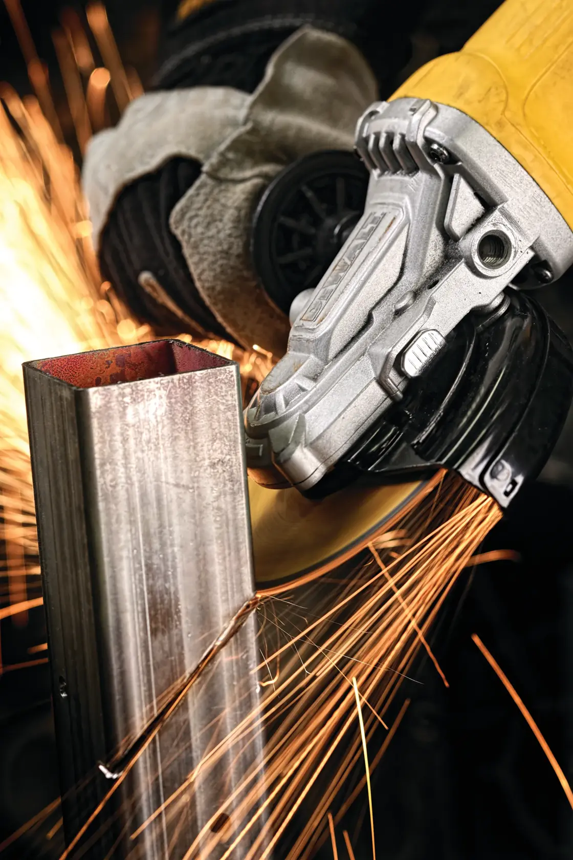 A close-up view of a DEWALT angle grinder cutting through a metal pipe, with sparks flying as the tool operates. The person using the grinder is wearing protective gloves.