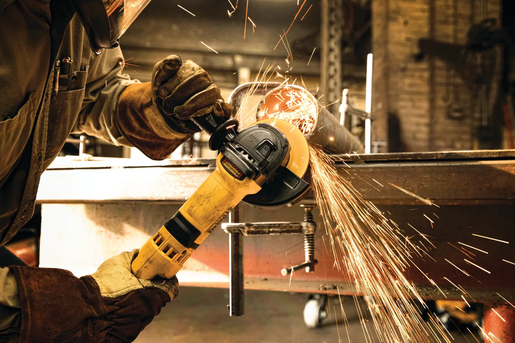 A person wearing protective gloves and a face shield is using a yellow DEWALT angle grinder to cut metal, producing sparks, in an industrial workshop setting.