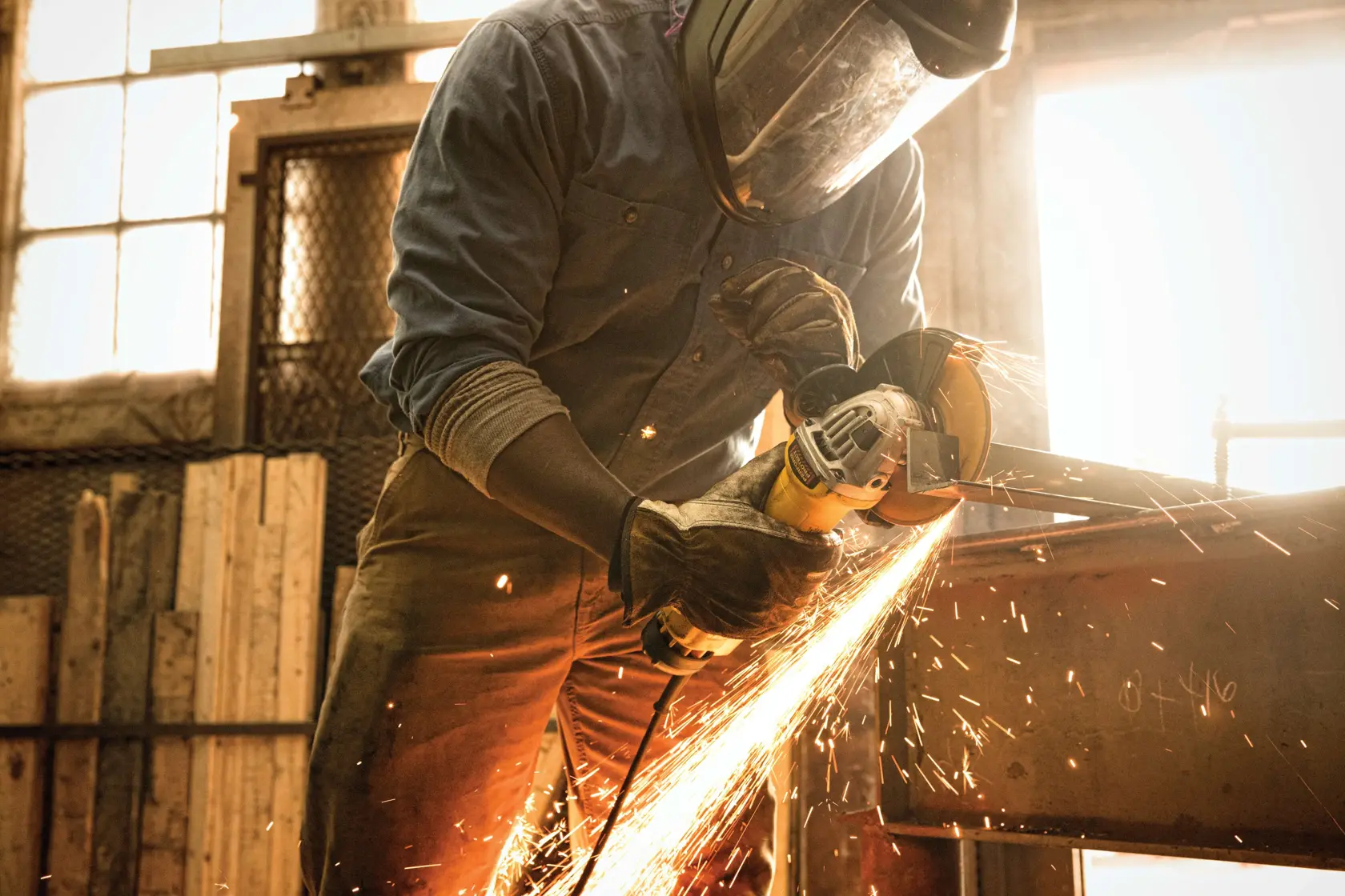 A person wearing protective gloves is using a DEWALT angle grinder to cut metal, with sparks flying in a workshop environment.