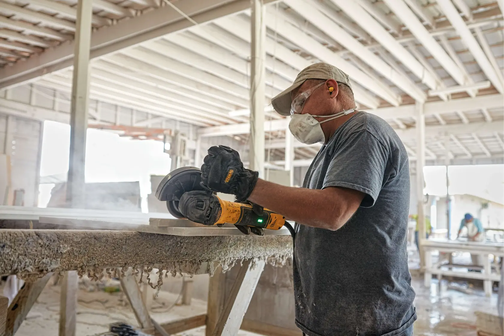 A person wearing protective gear, including gloves, a mask, and earplugs, is using a DEWALT angle grinder (model DWE43231VS) to cut or shape a slab in a workshop. The workspace appears dusty, and the person is working on a raised surface.