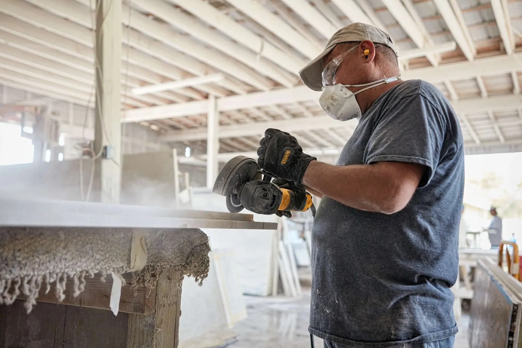 A person wearing a face mask, gloves, and a cap uses a DEWALT angle grinder to cut or sand a piece of material in an indoor workshop filled with dust.
