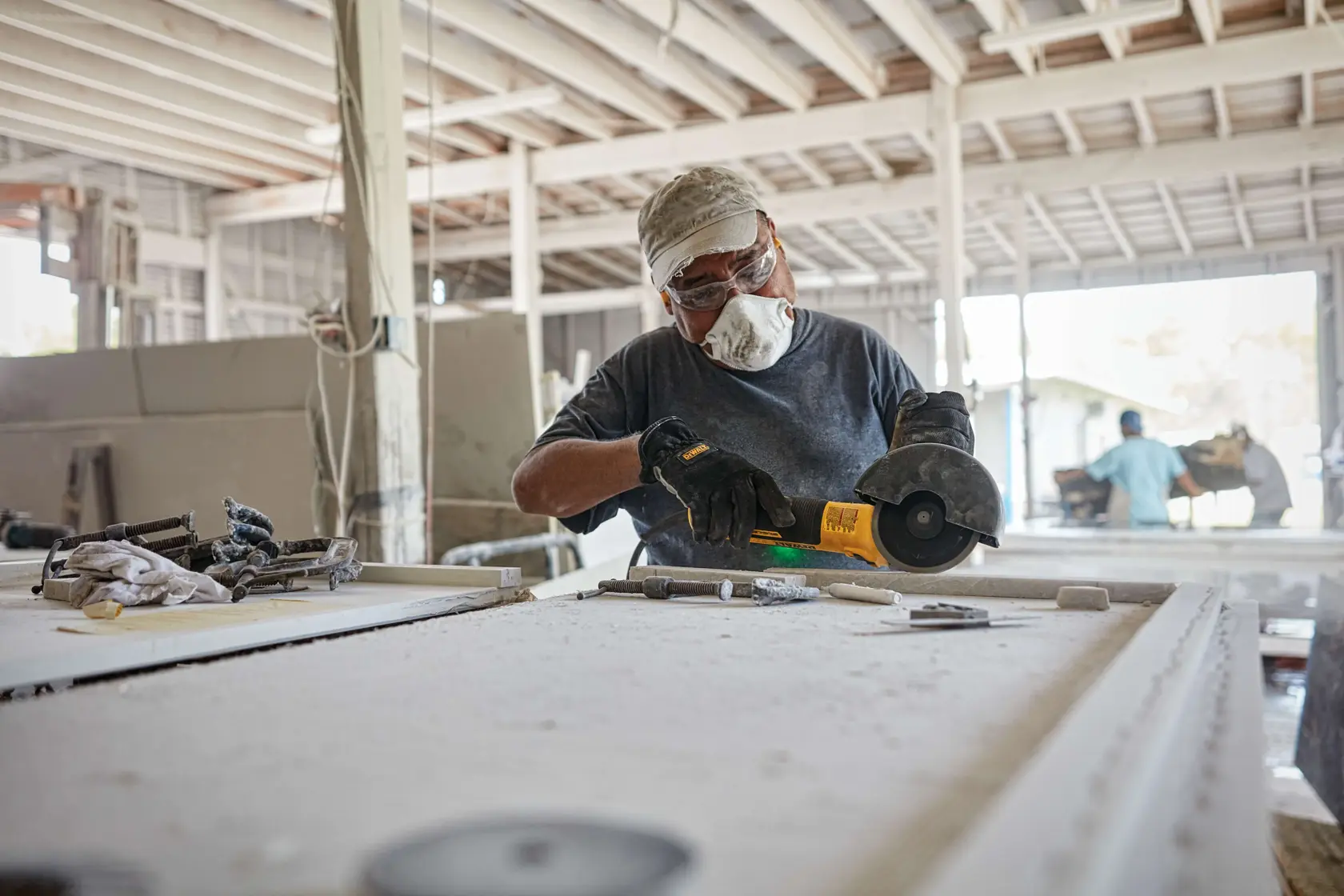 A person wearing gloves and a cap is using a yellow DEWALT angle grinder (model DWE43231VS) to cut or shape a material on a dusty workbench in an industrial workshop.