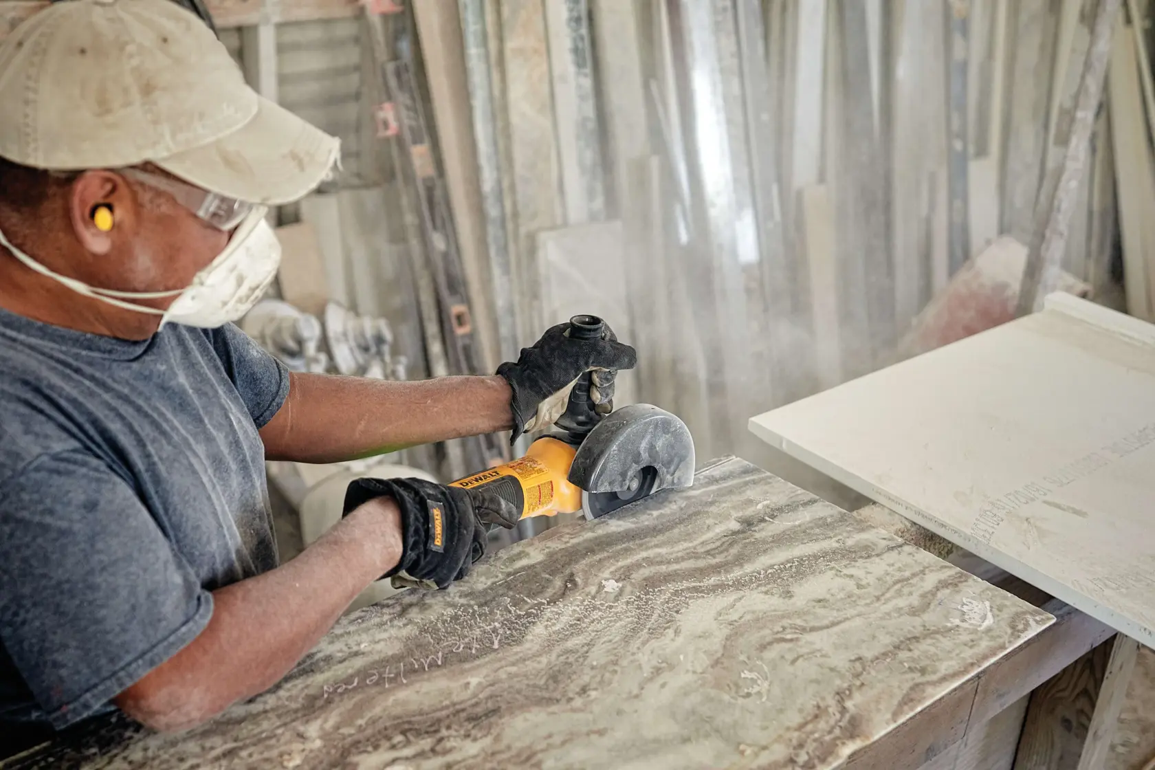 Person wearing gloves, ear protection, and face mask using a DEWALT angle grinder to cut or shape a stone or marble slab in a workshop.
