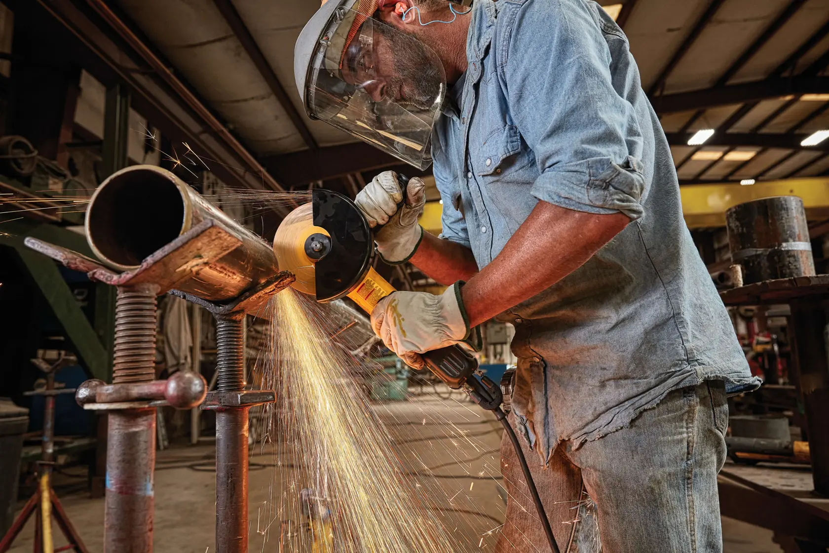 A person wearing protective gear uses a DEWALT angle grinder to cut a metal pipe in an industrial workshop, with sparks flying from the cutting area.