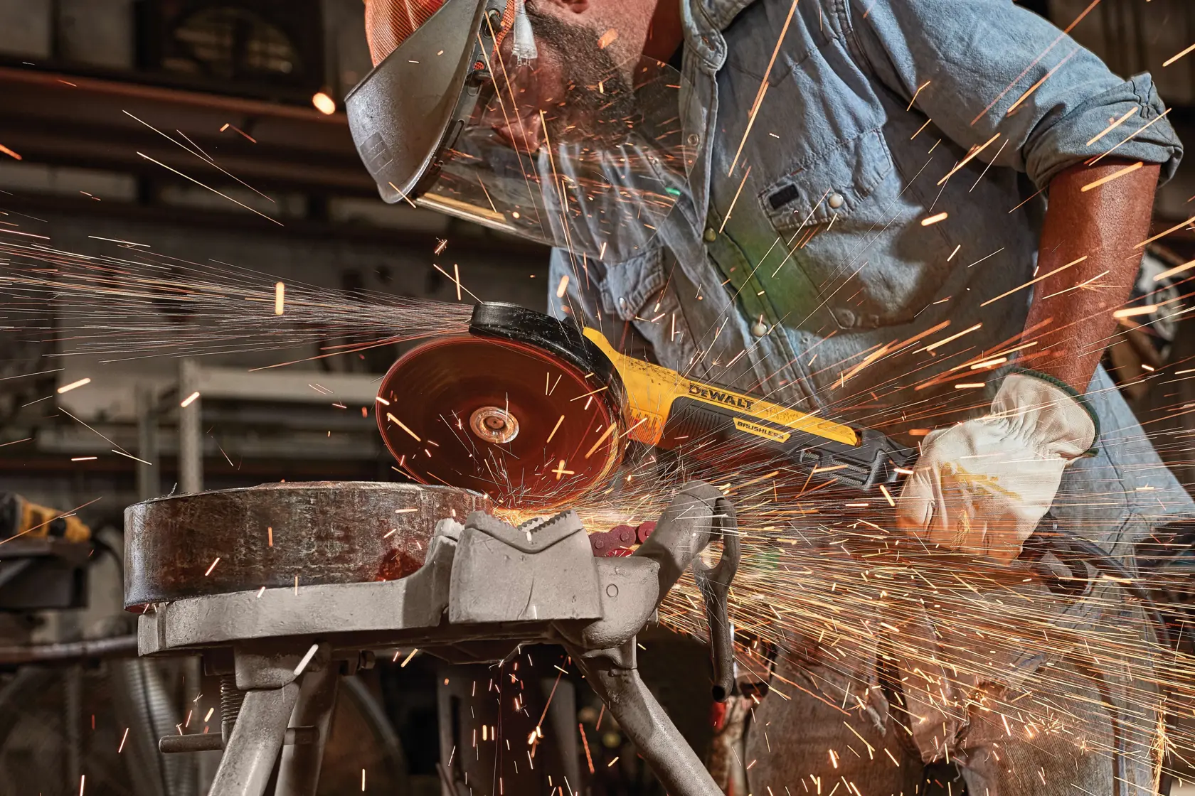 A person wearing protective gear uses a DEWALT angle grinder to cut metal, producing sparks, in an industrial workshop setting.