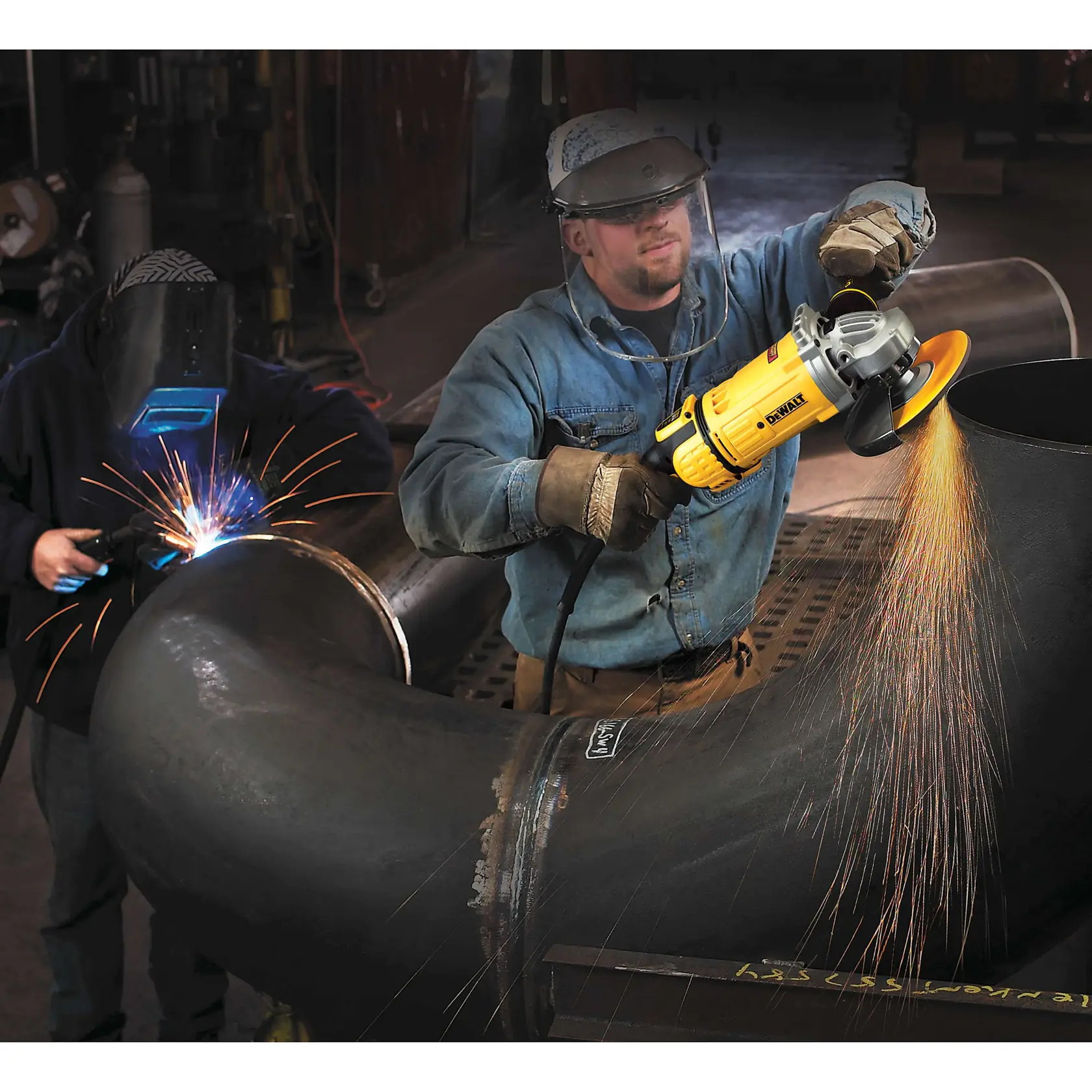 Two workers are operating in an industrial setting. One is using a DEWALT angle grinder, producing sparks on a large metal pipe, while the other is welding, with visible welding sparks and safety gear.