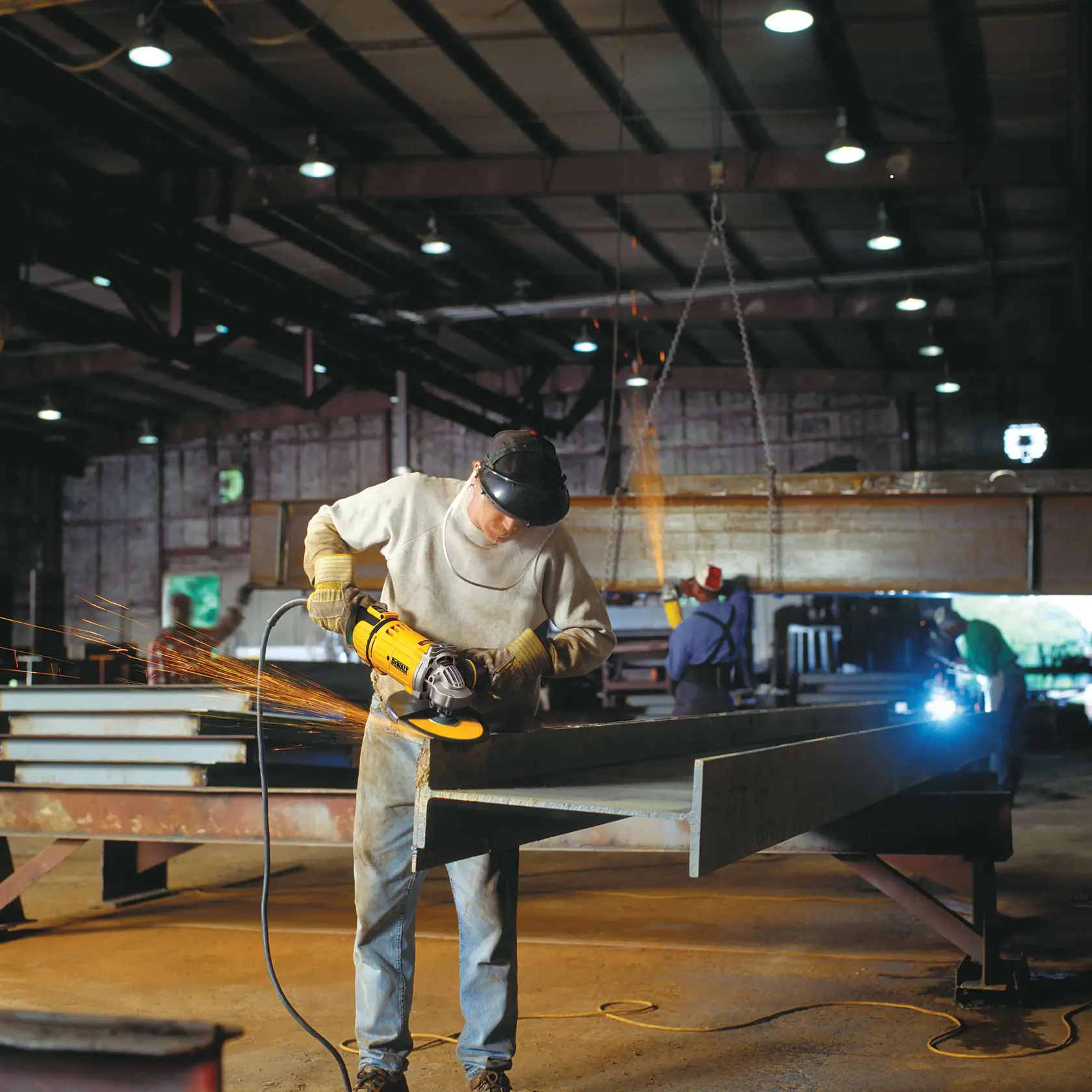 A person wearing protective gear is using a DEWALT angle grinder to cut or grind a large steel beam inside an industrial workshop. Sparks are flying from the metal. Other workers are seen in the background working with metal.
