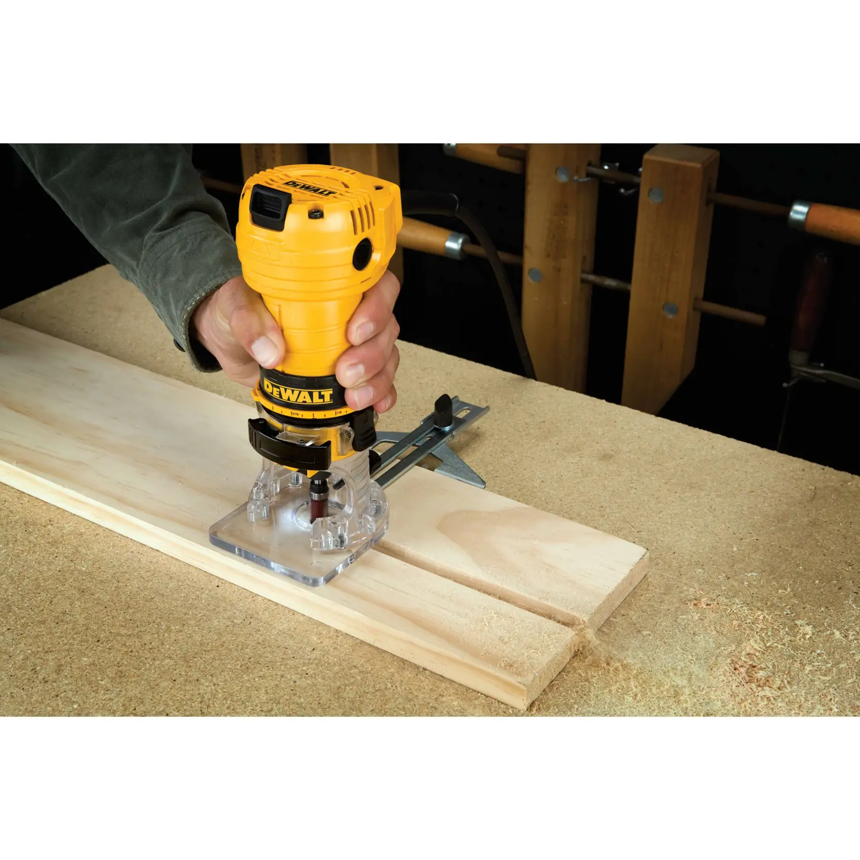 A person using a DEWALT DWE6000 woodworking router to carve or shape a piece of light-colored wood on a workbench. The router is yellow and black, and is positioned on top of the wood as sawdust collects around.