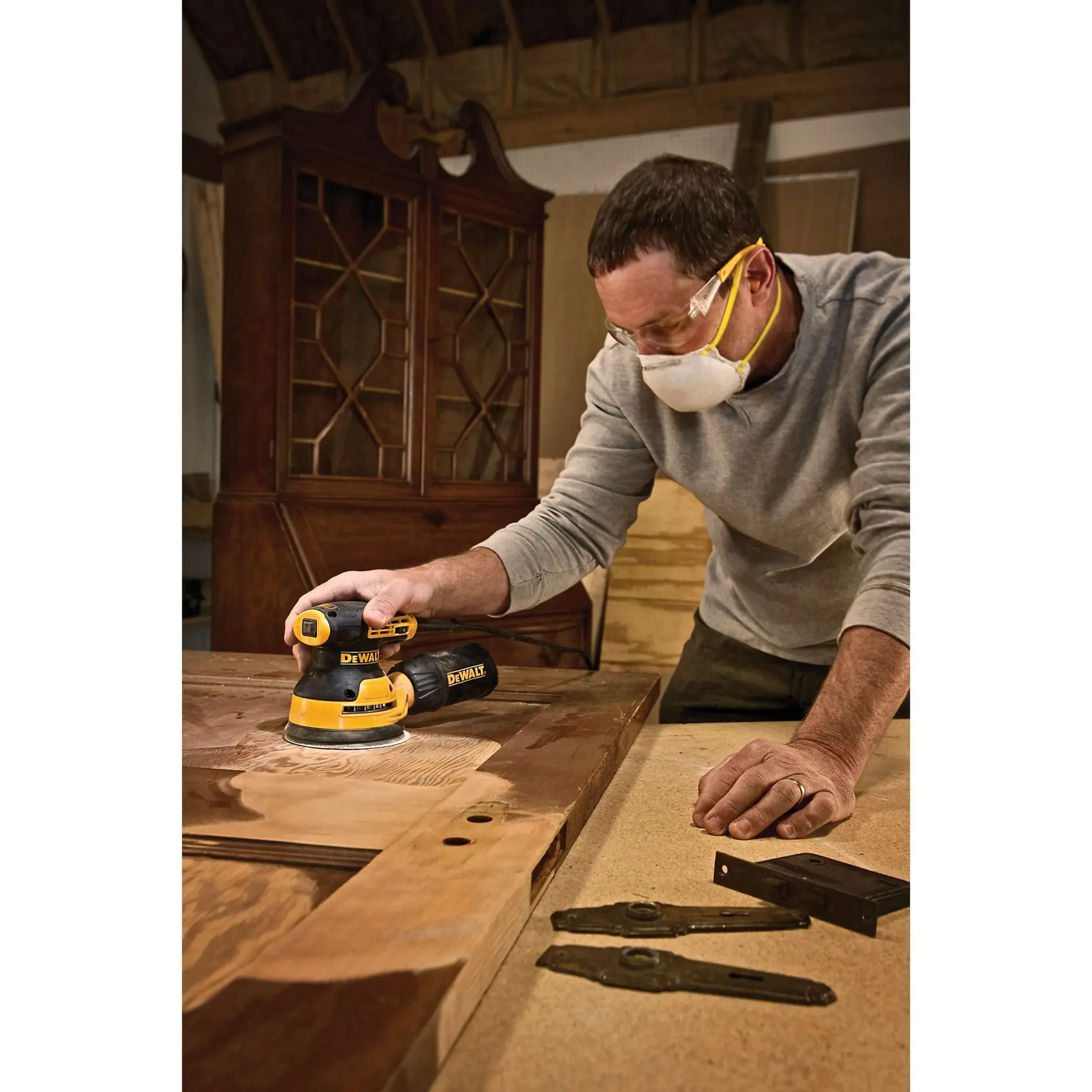 A person uses a DEWALT random orbital sander to sand a wooden door in a workshop. The individual is wearing a grey shirt and a dust mask, with antique cabinet hardware and another cabinet visible in the background.