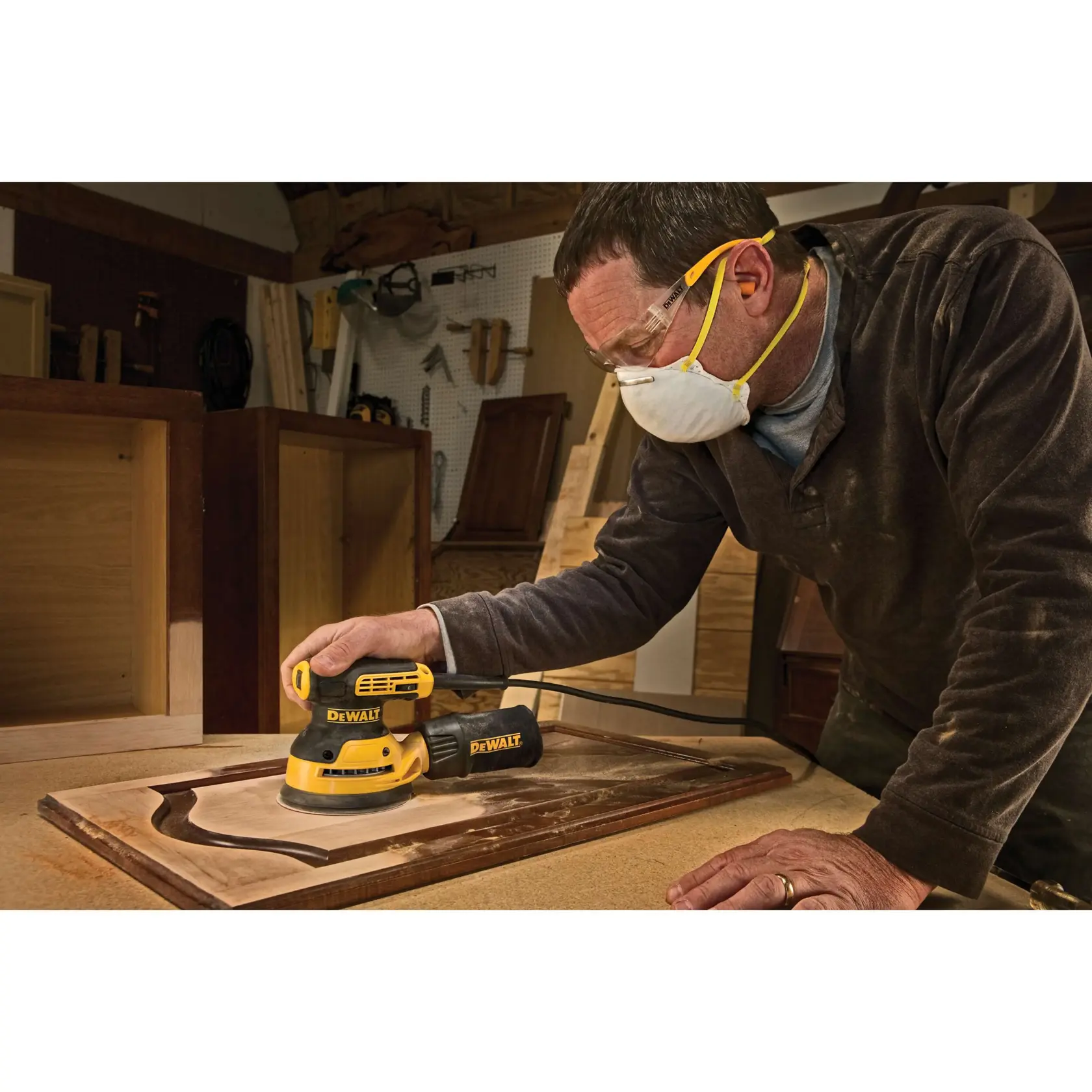 A person wearing safety goggles and a mask uses a DEWALT DWE6423K random orbit sander on a wooden surface in a workshop.