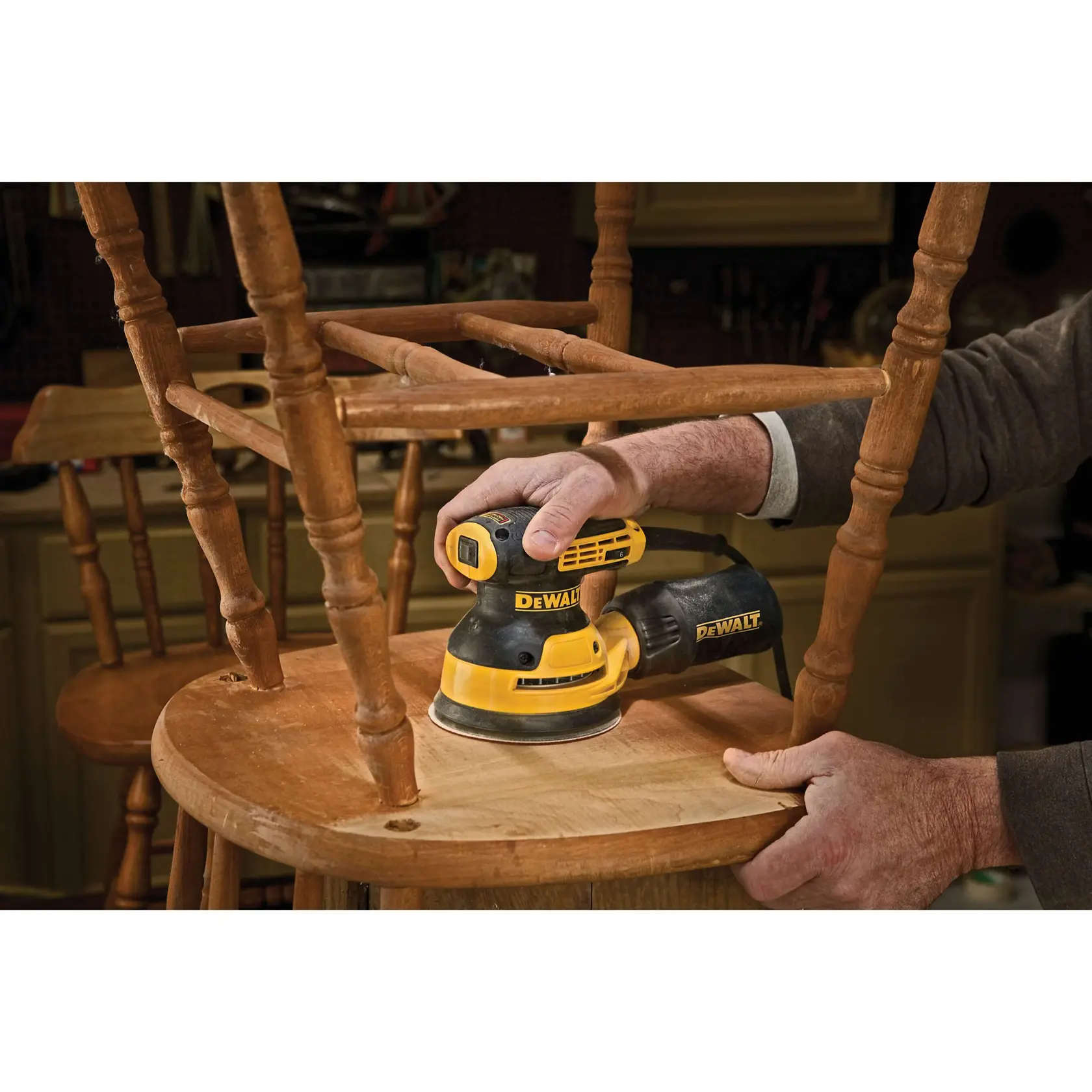 A person using a DEWALT DWE6423K random orbit sander to sand the seat surface of a wooden chair in a workshop.