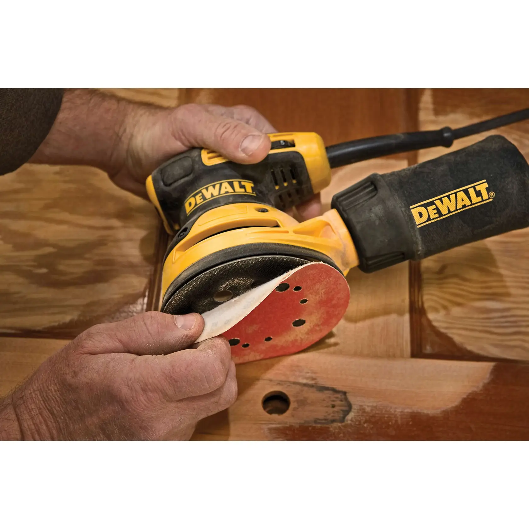 A DEWALT random orbit sander is being held by hands, with a sanding disc being attached or removed. The sander is yellow and black, and is on a wooden surface.