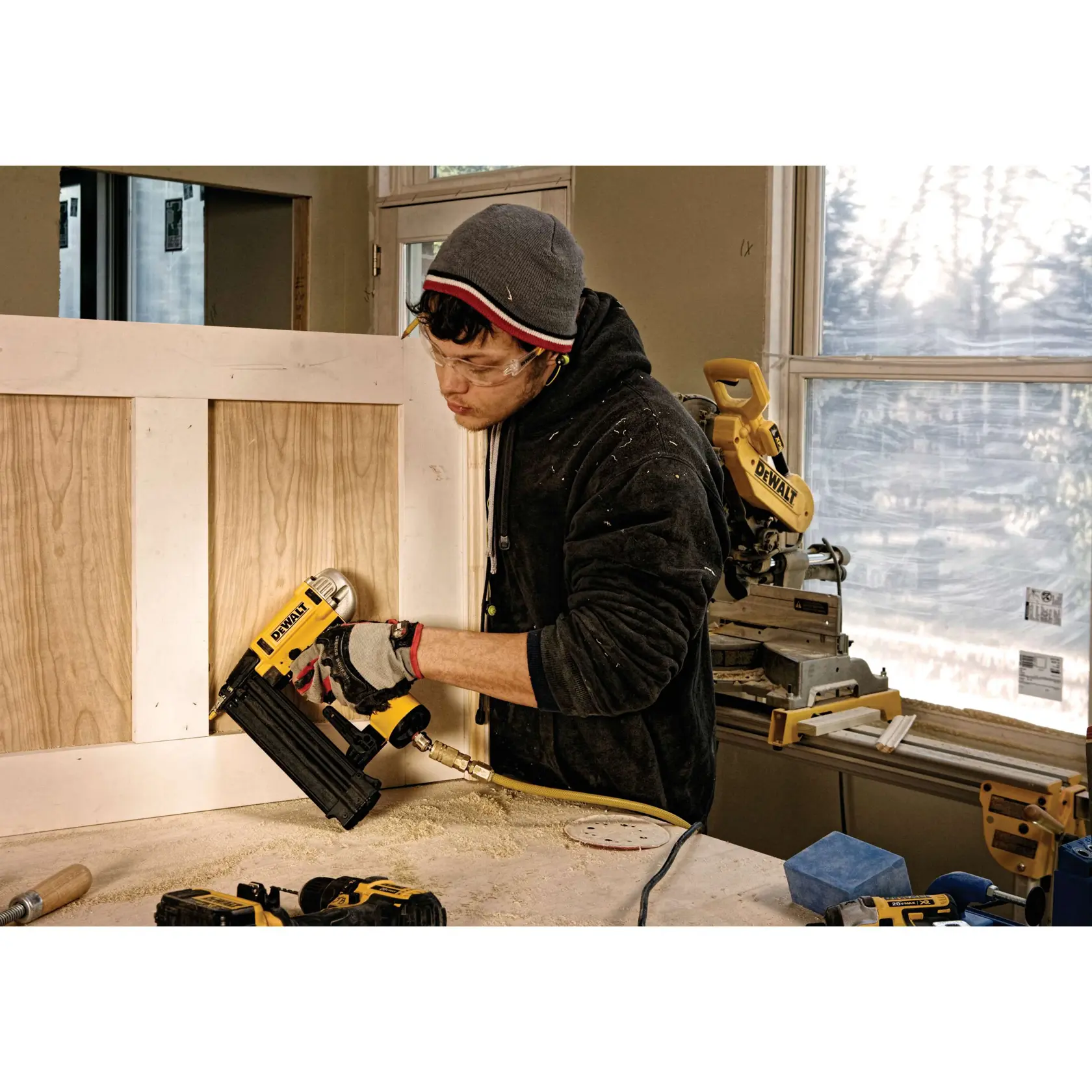 A person wearing a beanie and gloves is using a DEWALT DWFP12233 pneumatic nailer on a wooden panel in a workshop. Various DEWALT tools are visible, including a miter saw, with sunlight streaming through a window in the background.