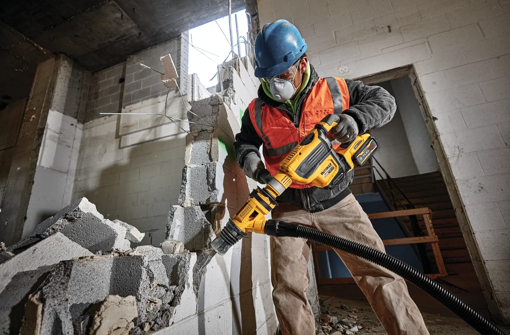 A construction worker wearing a blue helmet and orange safety vest uses a yellow DEWALT power tool equipped with a dust extraction attachment to demolish a concrete wall in an unfinished building.