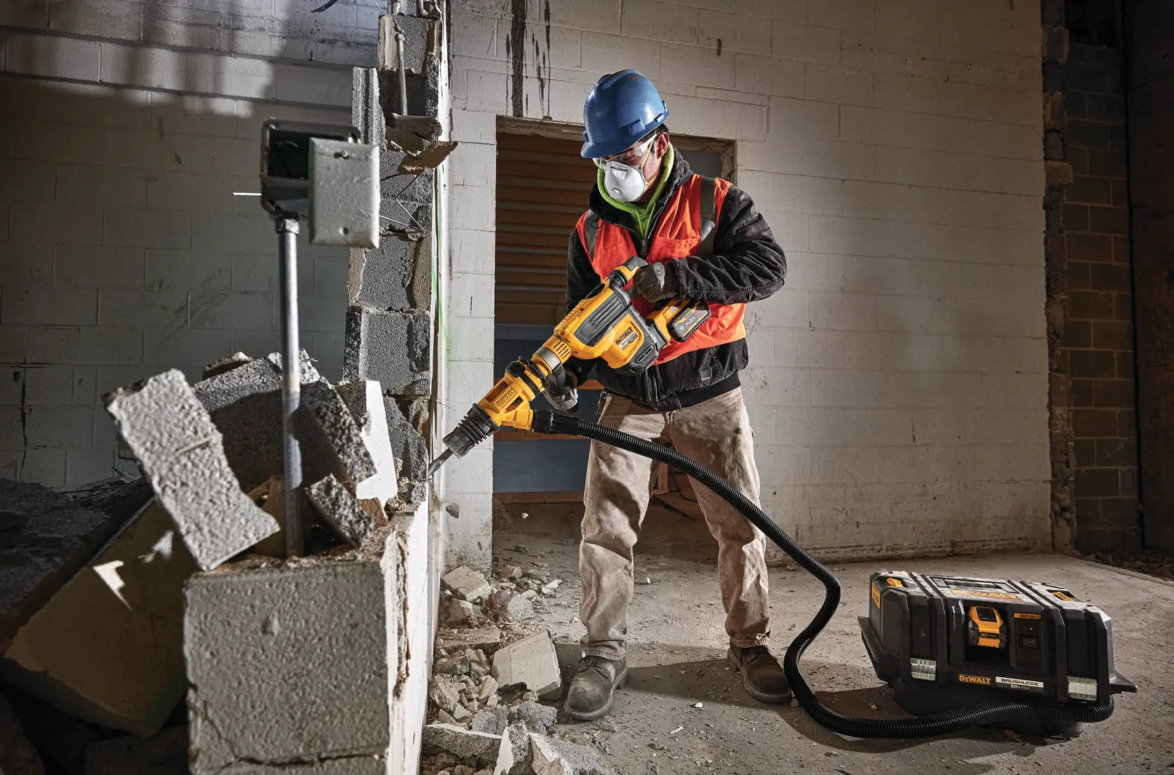 A construction worker wearing a blue hard hat and orange safety vest uses a yellow DEWALT demolition tool with an attached hose to break apart a concrete wall. Rubble is scattered on the floor and a DEWALT equipment case sits nearby.