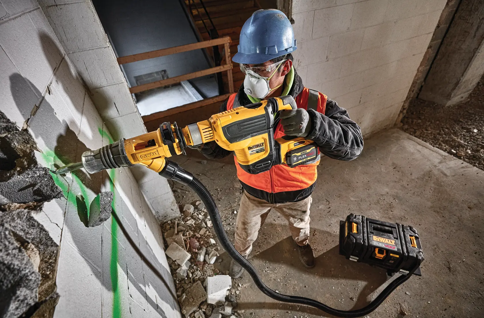 A construction worker wearing a blue hard hat and orange safety vest is using a yellow DEWALT rotary hammer drill with a dust extraction hose to drill into a concrete wall. There is construction debris on the floor and a DEWALT tool case nearby.