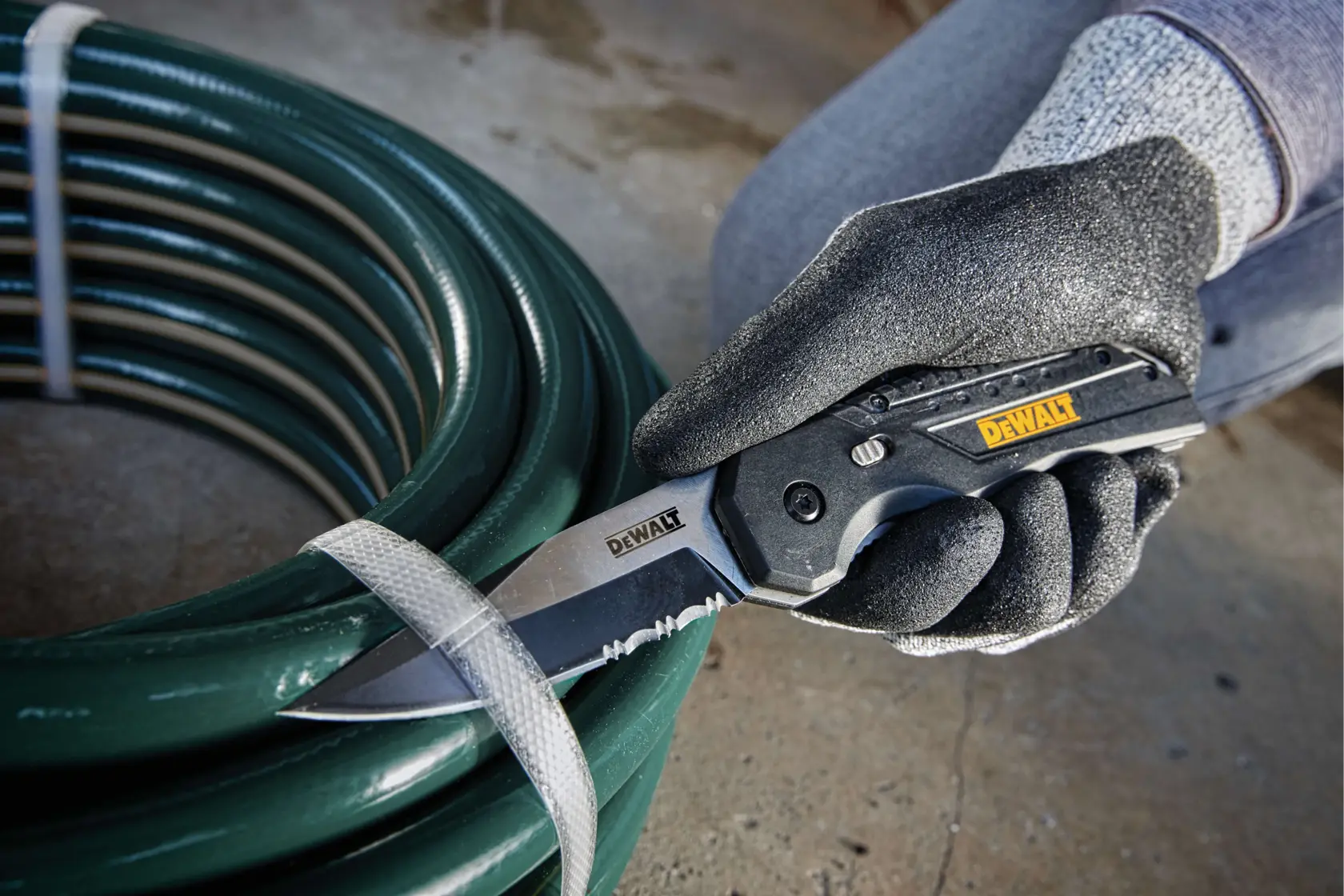 A gloved hand is using a DEWALT folding knife to cut a strap securing a coil of green hose on a concrete floor.