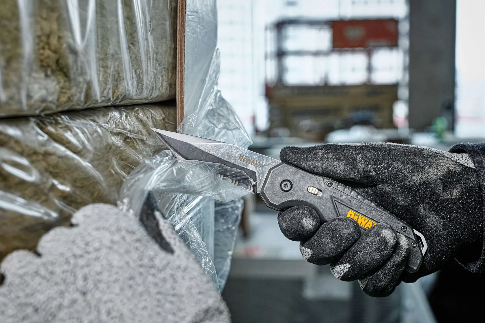 A close-up of a gloved hand using a DEWALT folding utility knife to cut plastic wrapping on a package, with another gloved hand holding the material in place. The scene appears to be in an industrial or construction setting.