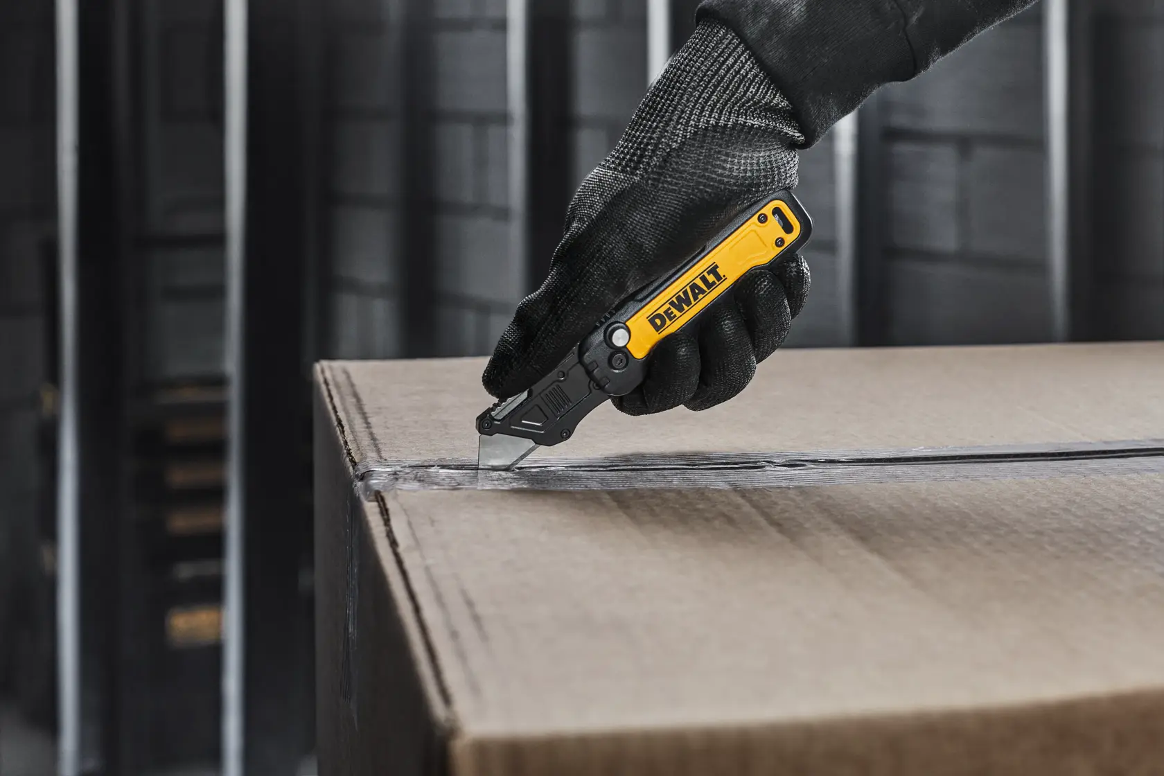A person wearing a black glove uses a yellow and black DEWALT utility knife to cut open a cardboard box.