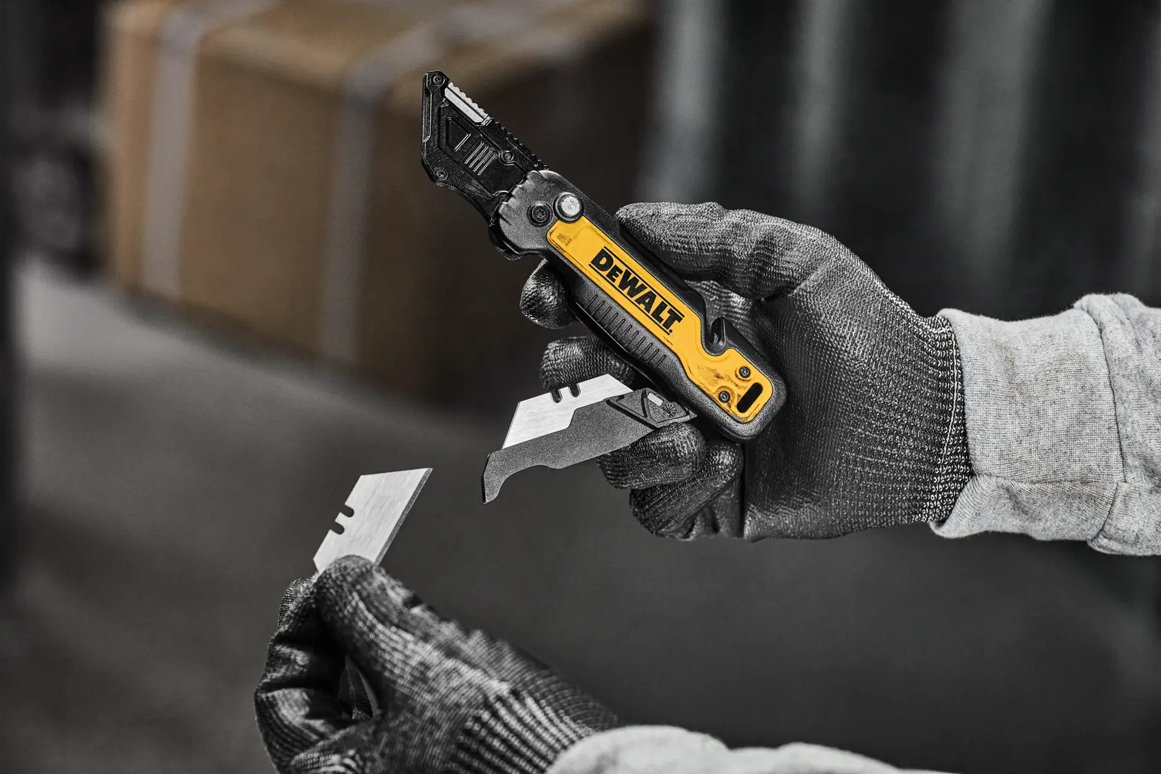 A person wearing work gloves is holding a DEWALT utility knife and a replacement blade, preparing to insert the blade into the tool.